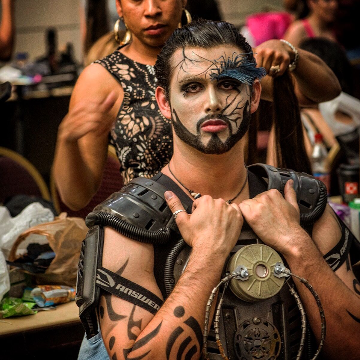 A man with face paint and tattooed arms wears a black shoulder harness with a round metal plate at the chest while someone behind him works on another model's hair.