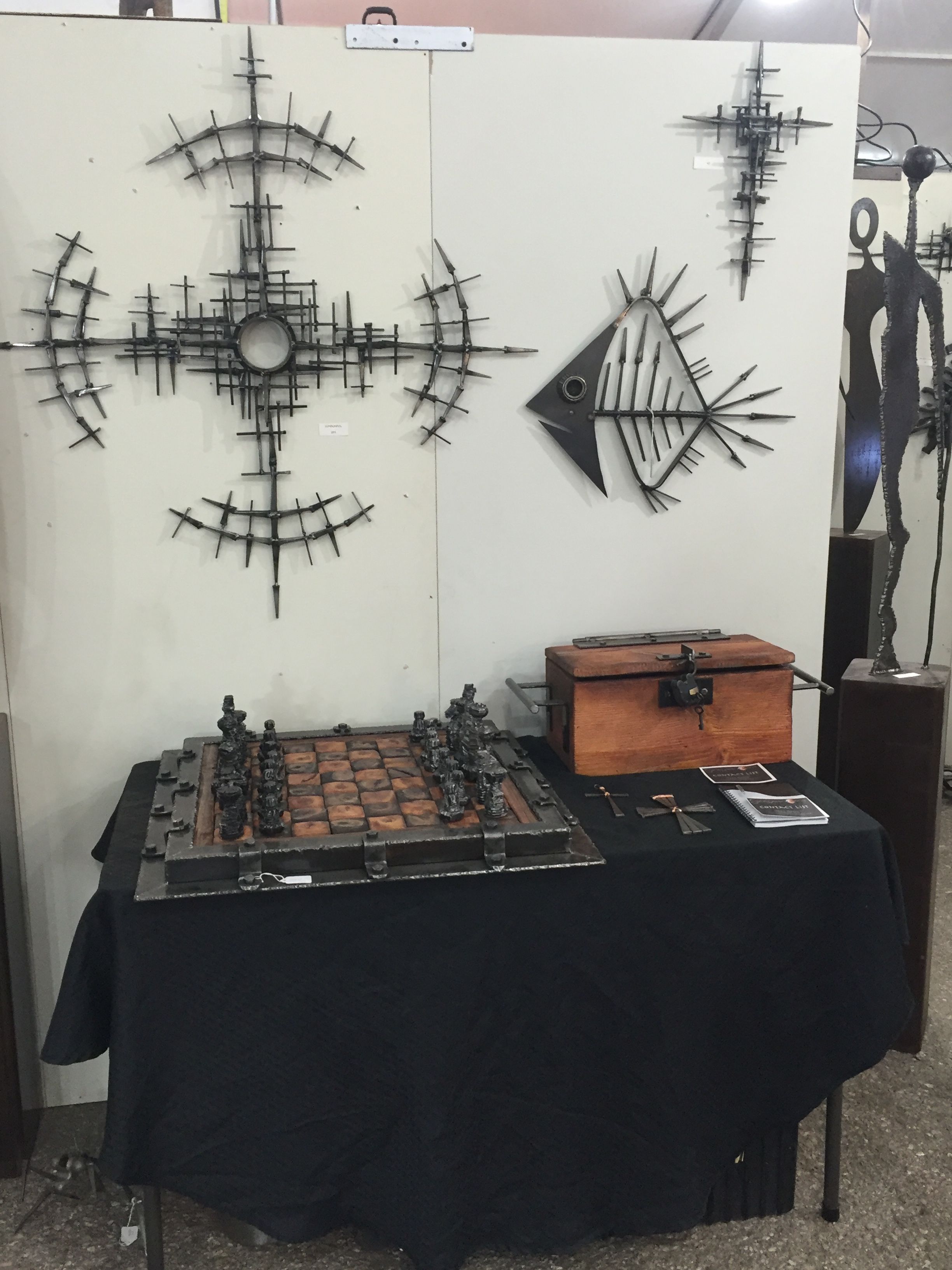 A chess set and a wooden box on a black table, below a steel sunburst and a fish skeleton on a white wall.