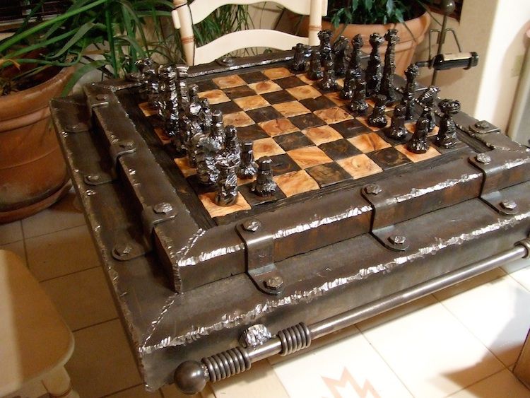A chess set on a thick wooden board edged in steel, with a rolled steel bar along its front edge.