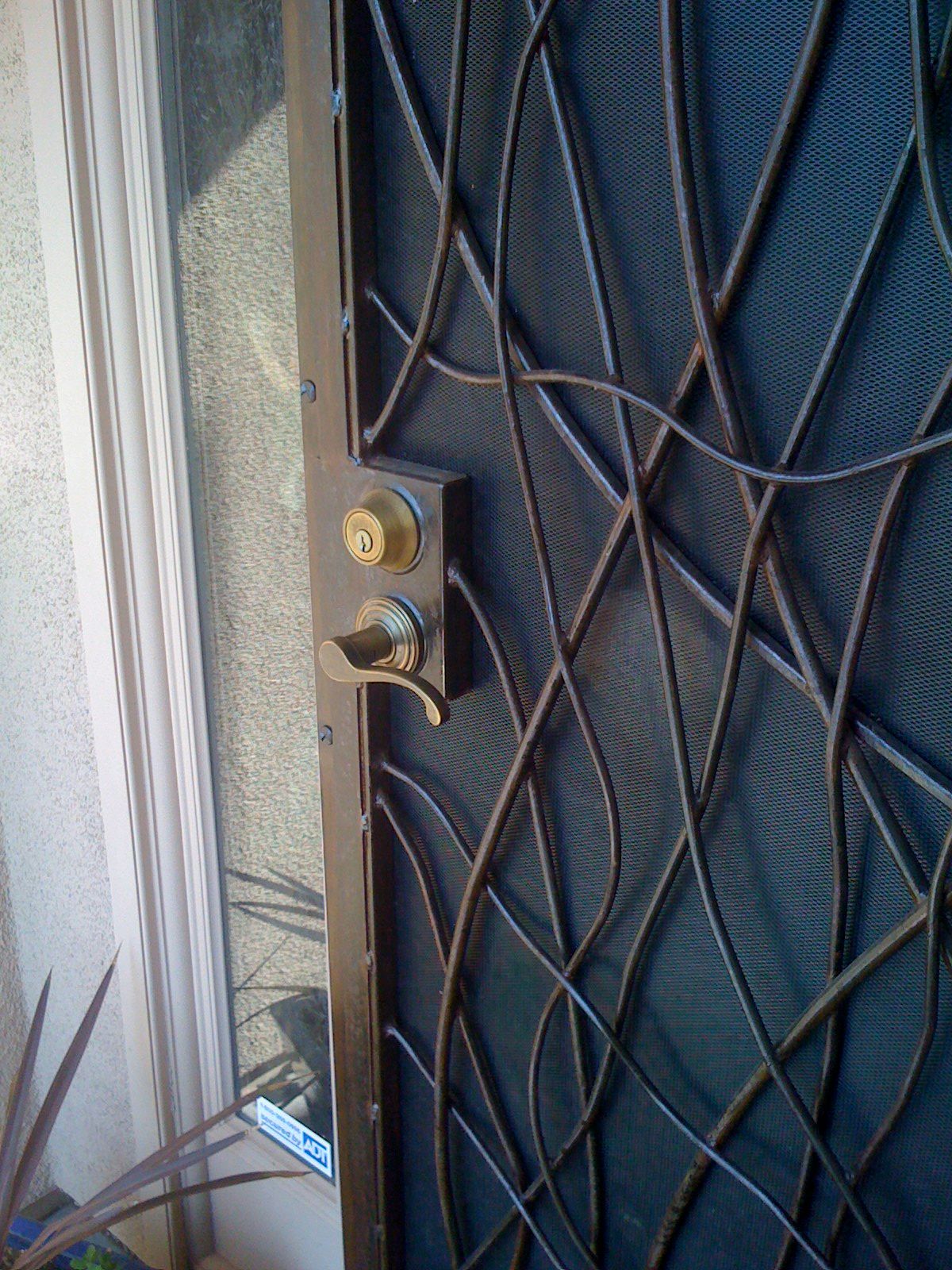 A dark security door filled with long curving and crossing lines, beside a window, with a brass lock.