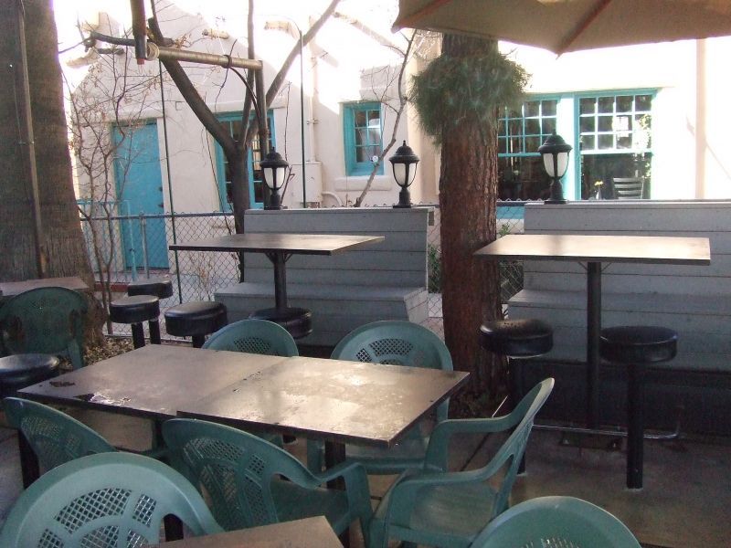 Square metal-topped tables and green plastic chairs on a restaurant patio, with a tree trunk and a stucco wall behind.
