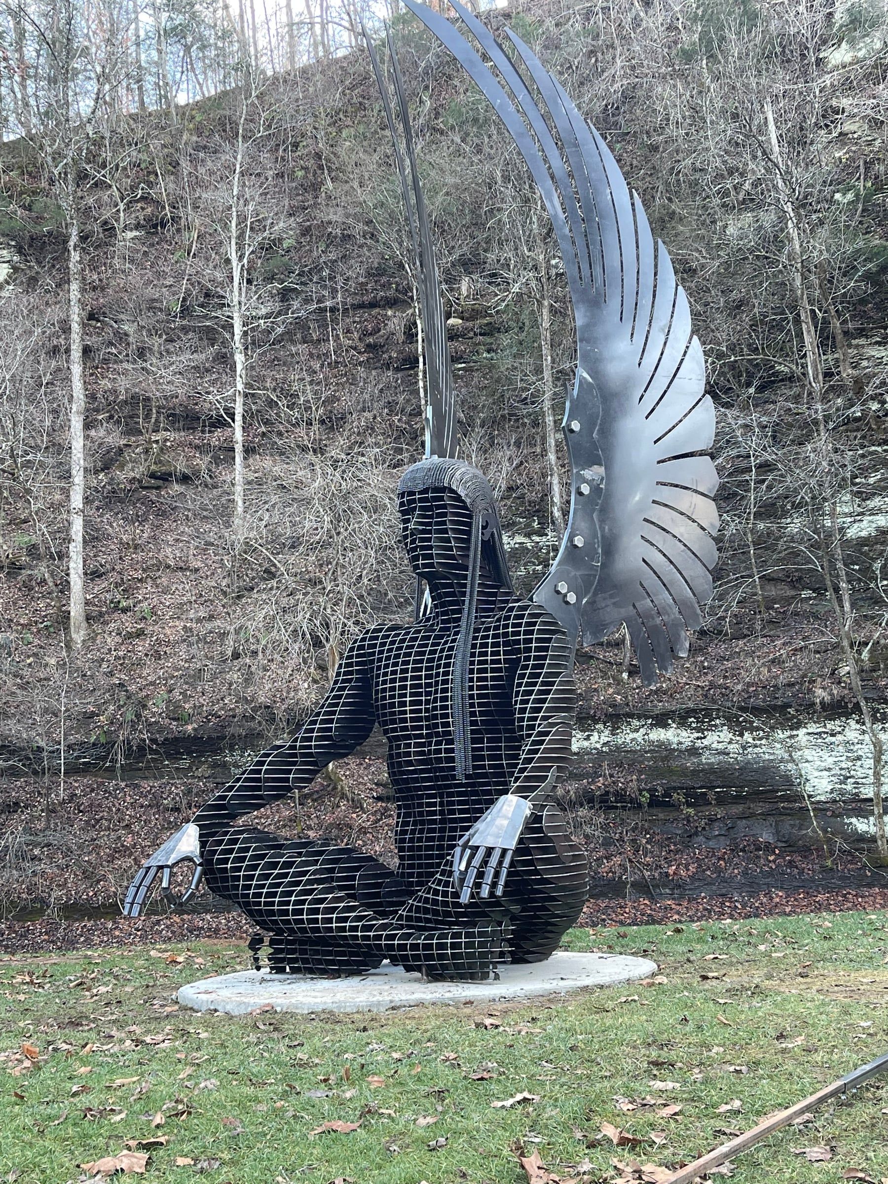 A cross-legged steel angel in meditation, hands resting on her knees and tall wings rising behind her, on a lawn before a rock bluff and bare trees.