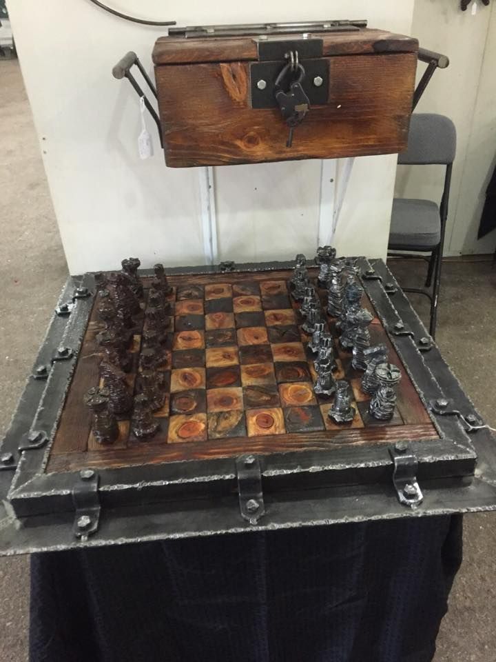 A chess set in a riveted steel frame on a black-draped table, with a wooden box with a steel latch and handles above it.