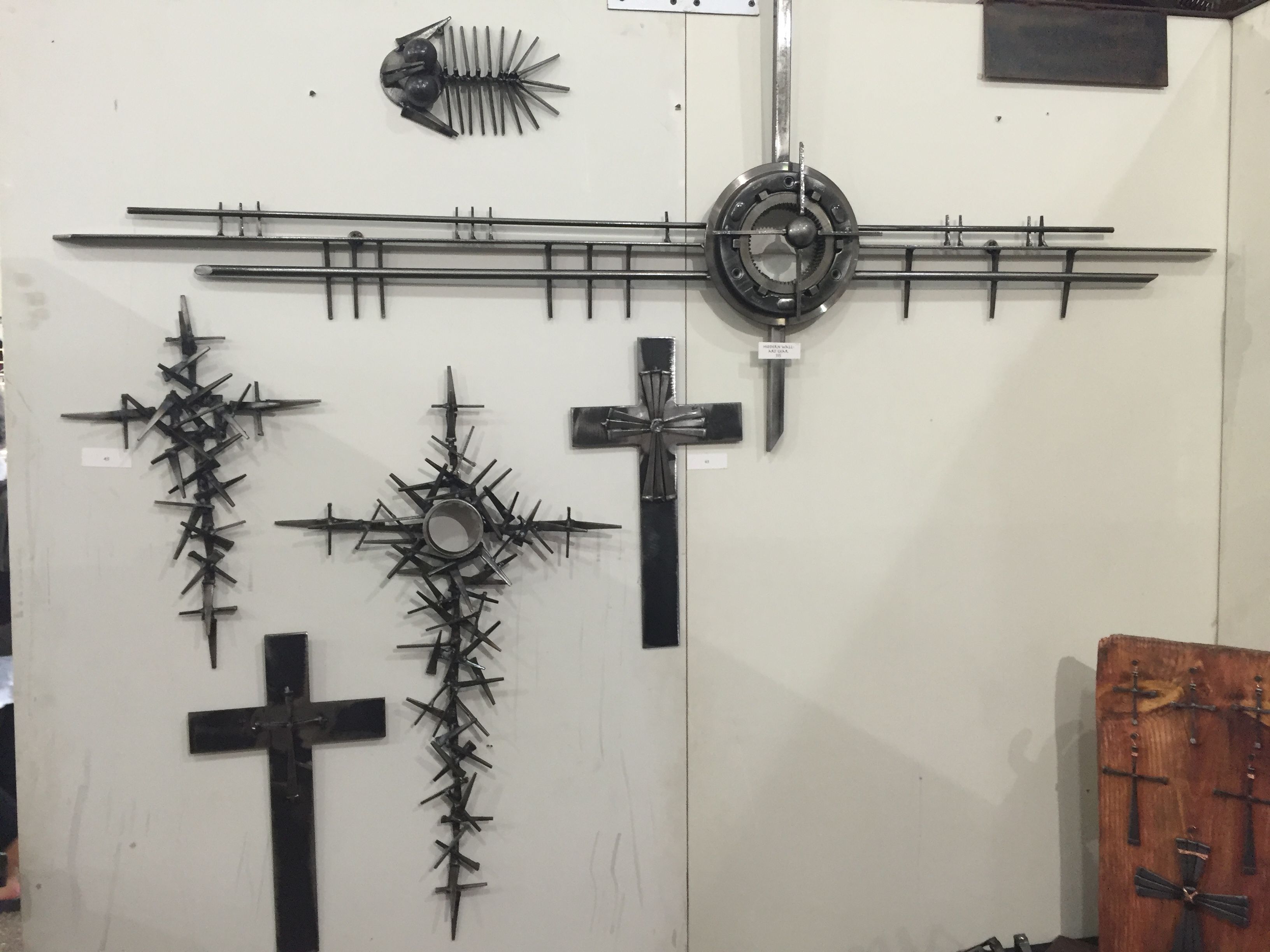 Several black steel crosses of different shapes and a long horizontal piece with a ring, hung on a white wall.