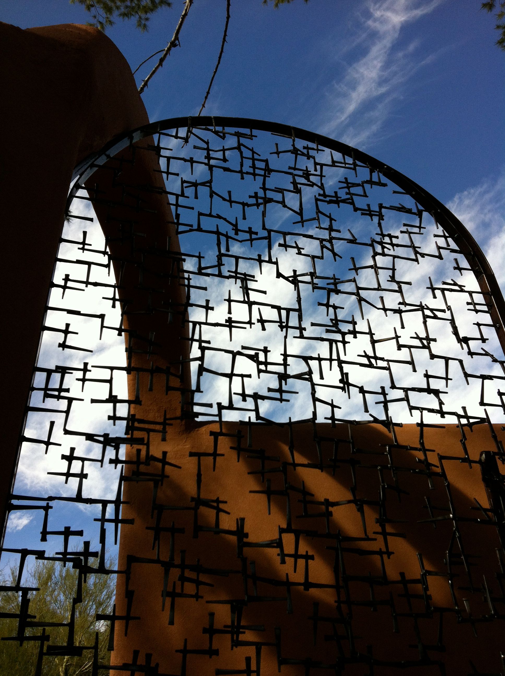 Looking up at the arched top of the nail gate, its lattice dark against a blue sky with thin clouds, beside the curve of an adobe wall.