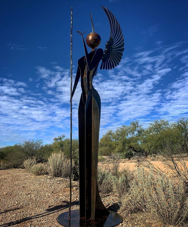 A tall, slender dark steel angel with a fanned wing and a staff topped by a sphere, standing in the desert under a blue sky.