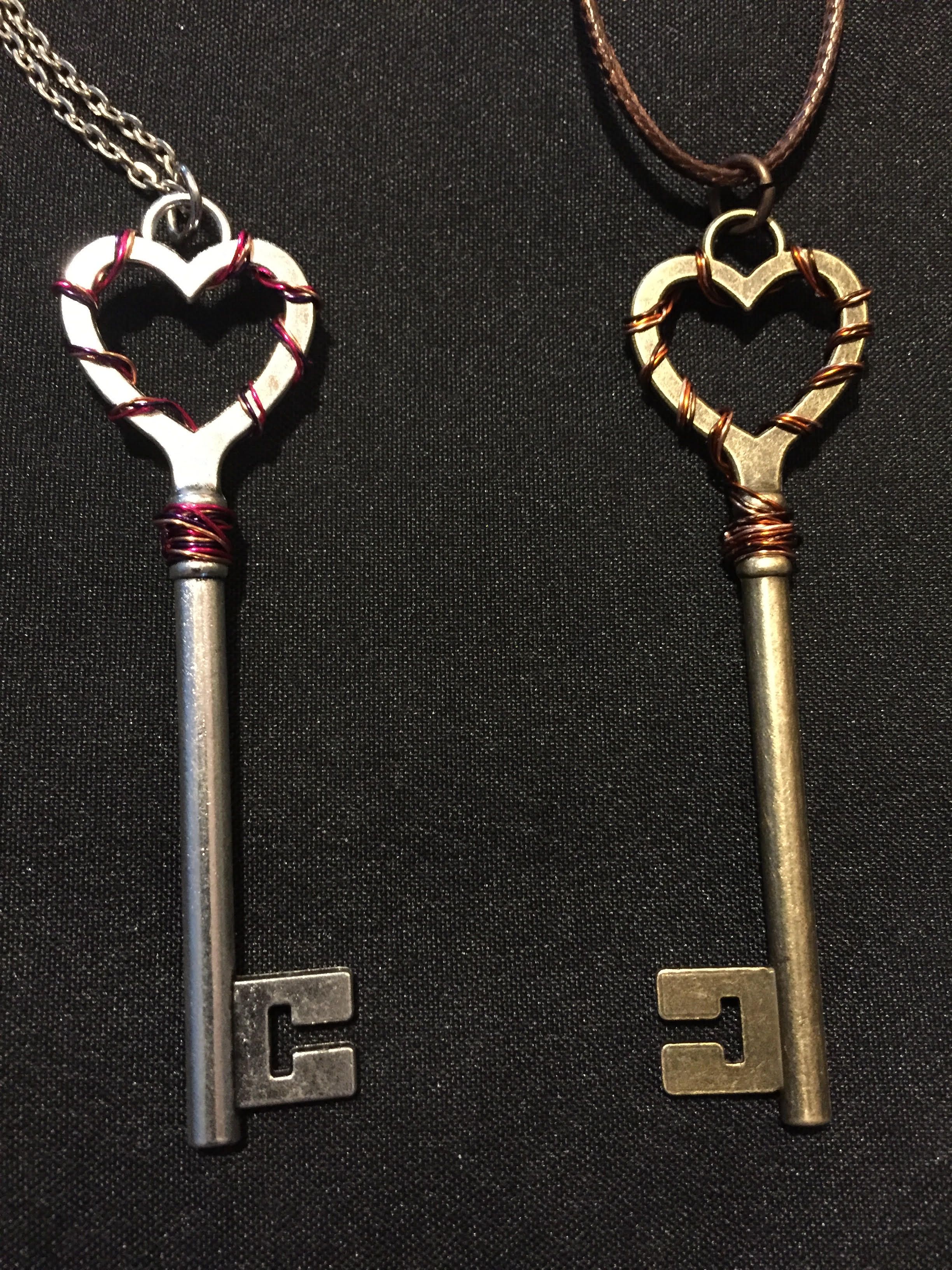 Two long key pendants with heart-shaped bows, one silver and one brass, wrapped with red and copper wire, on black cloth.