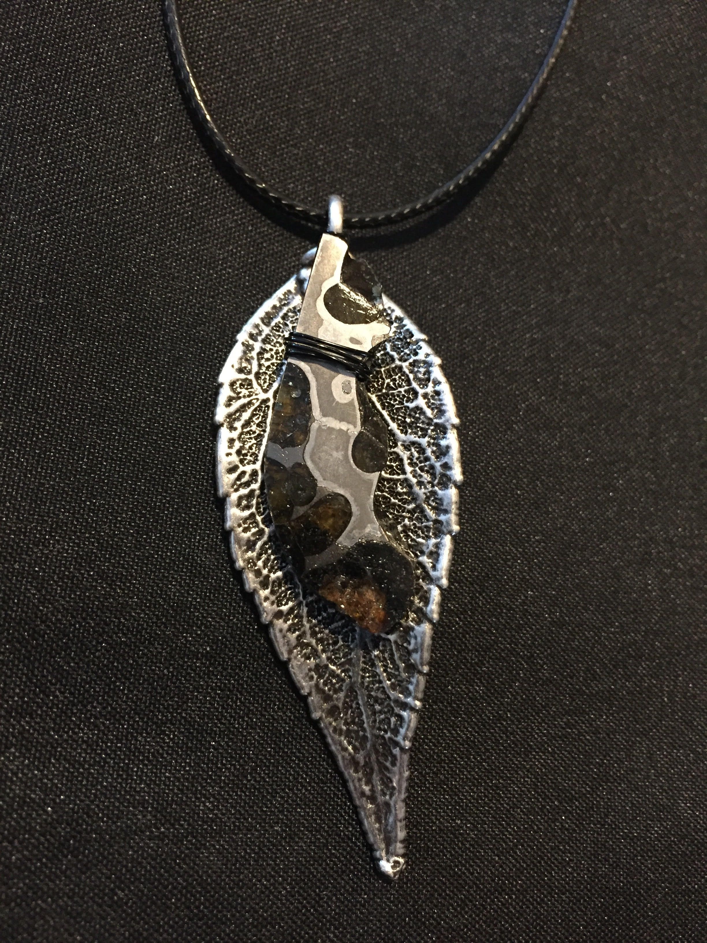A silver leaf-skeleton pendant with a dark polished inlay, on a black cord against black cloth.