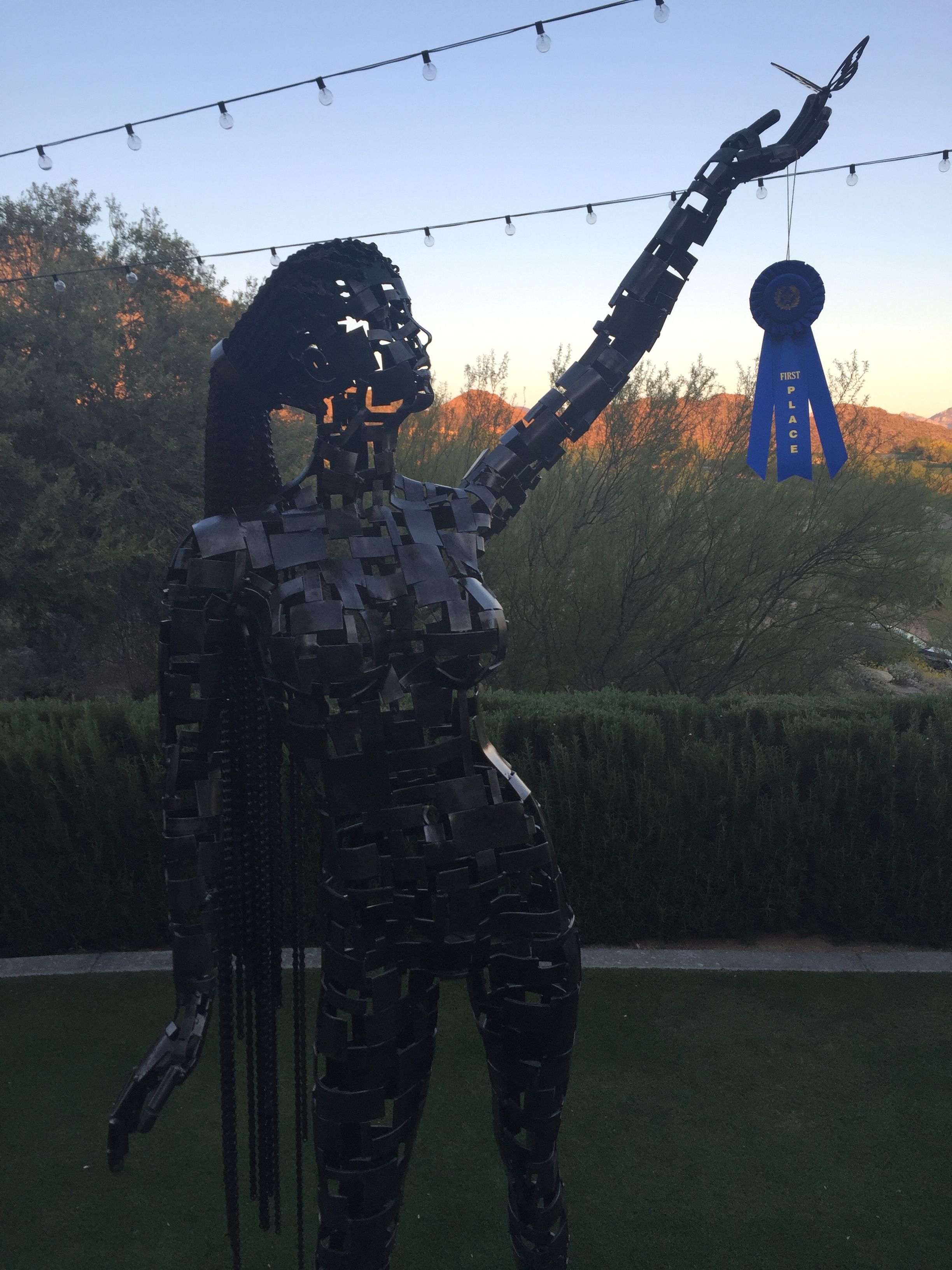 A standing steel figure of a woman with one arm raised, a blue ribbon hanging from her hand, against a dusk sky with string lights and desert hills.