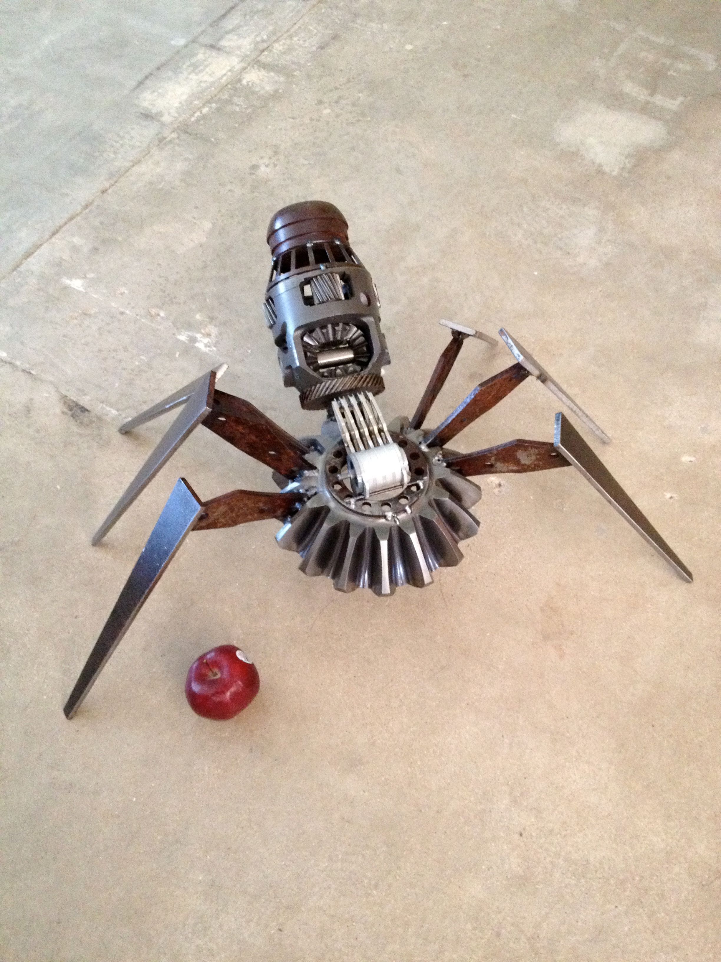 A steel spider on a concrete floor, its body built around a gear and a ribbed cylinder, standing on long pointed legs cut from steel plate.