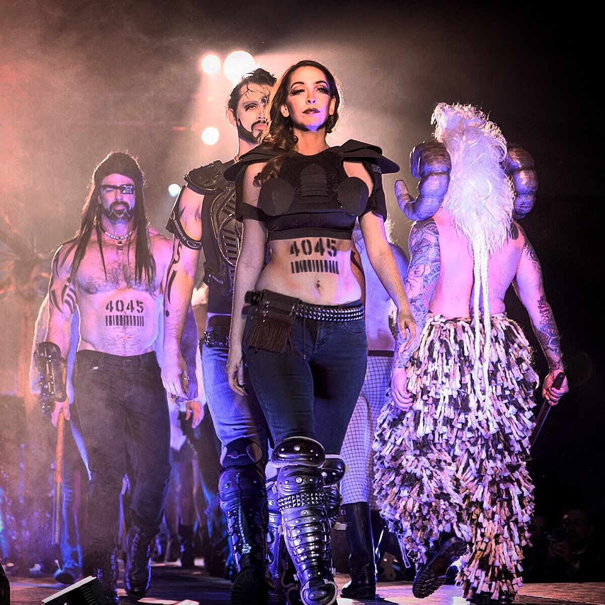 Models walk a smoky runway toward the camera; the woman in front wears a black armored top with 4045 painted on her stomach, and a white feathered costume follows on the right.