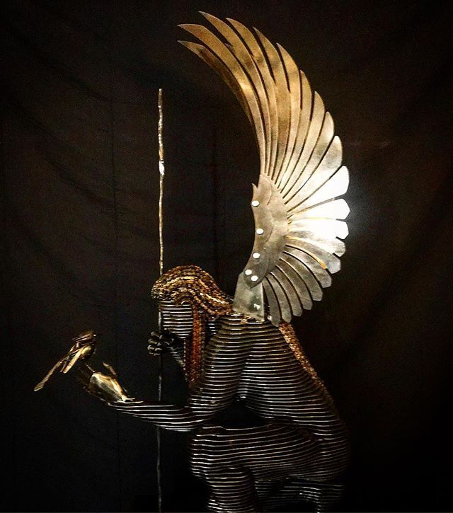 A kneeling angel with a golden fanned wing and gold-banded hair, one hand held out with a small gold piece in it and a spear behind, lit against a dark background.