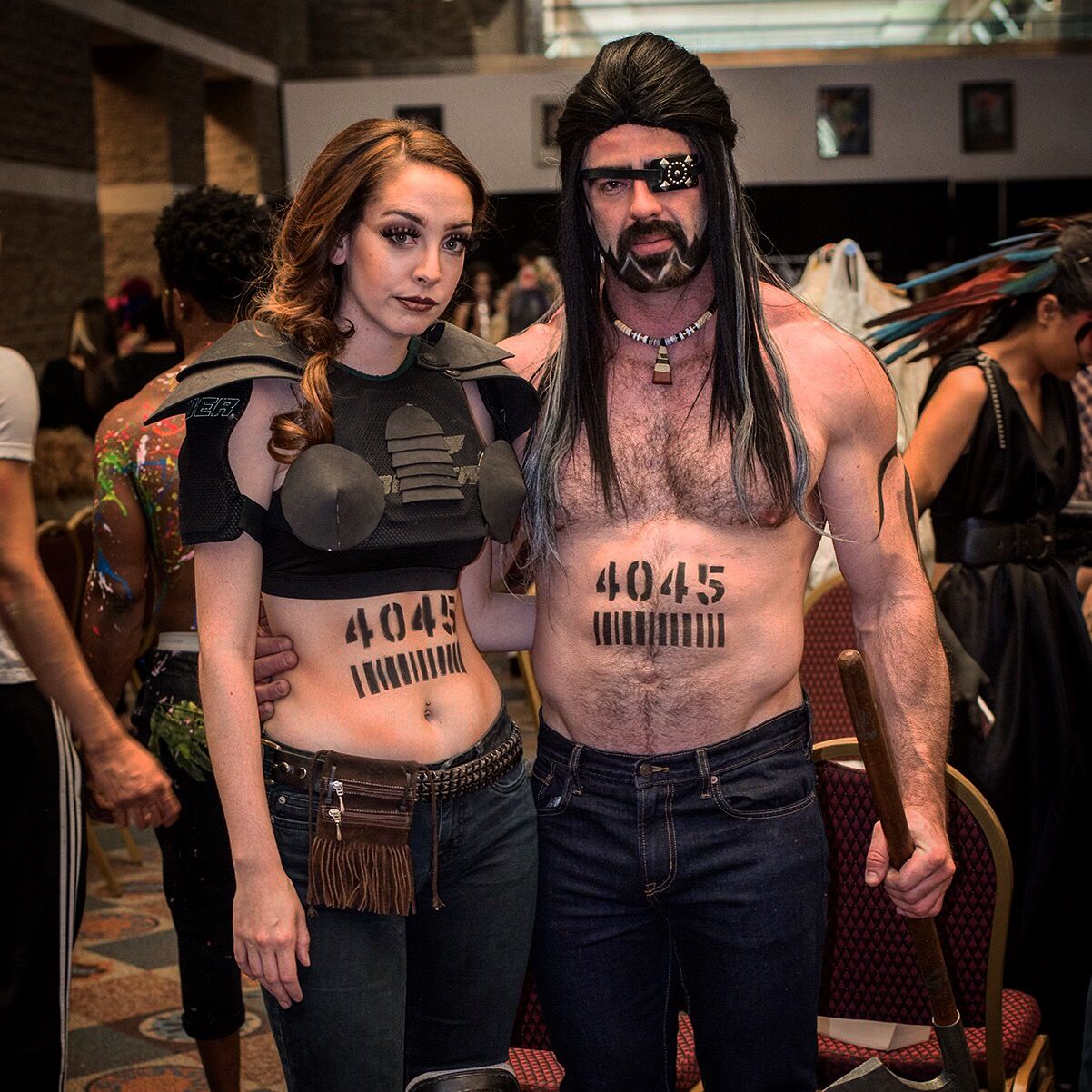 A woman and a man stand side by side in a dressing room, each with 4045 and a barcode painted on the stomach; he wears an eyepatch and long dark hair.