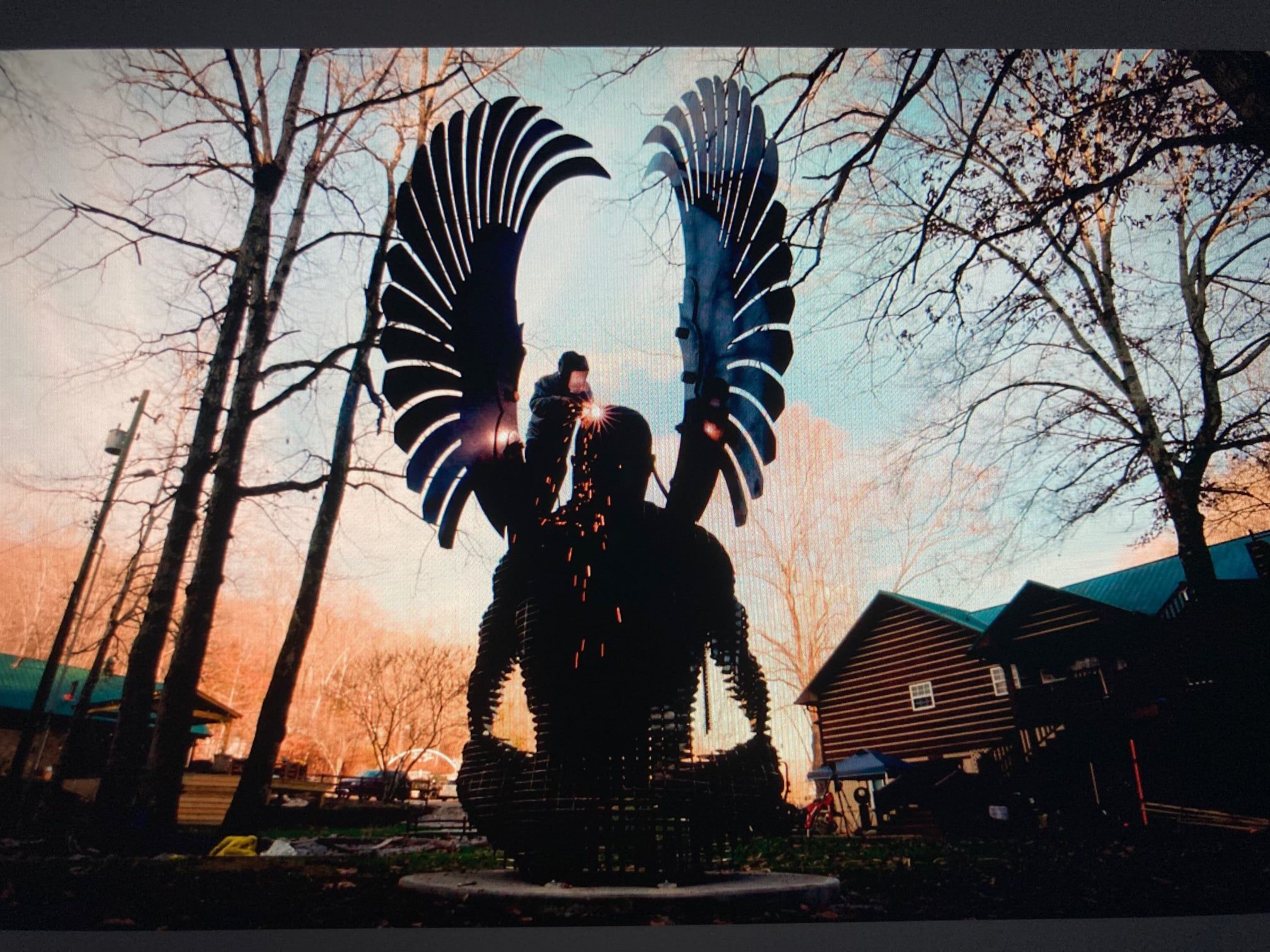 The silhouette of a seated winged figure against an orange dusk sky, among bare trees and cabins.