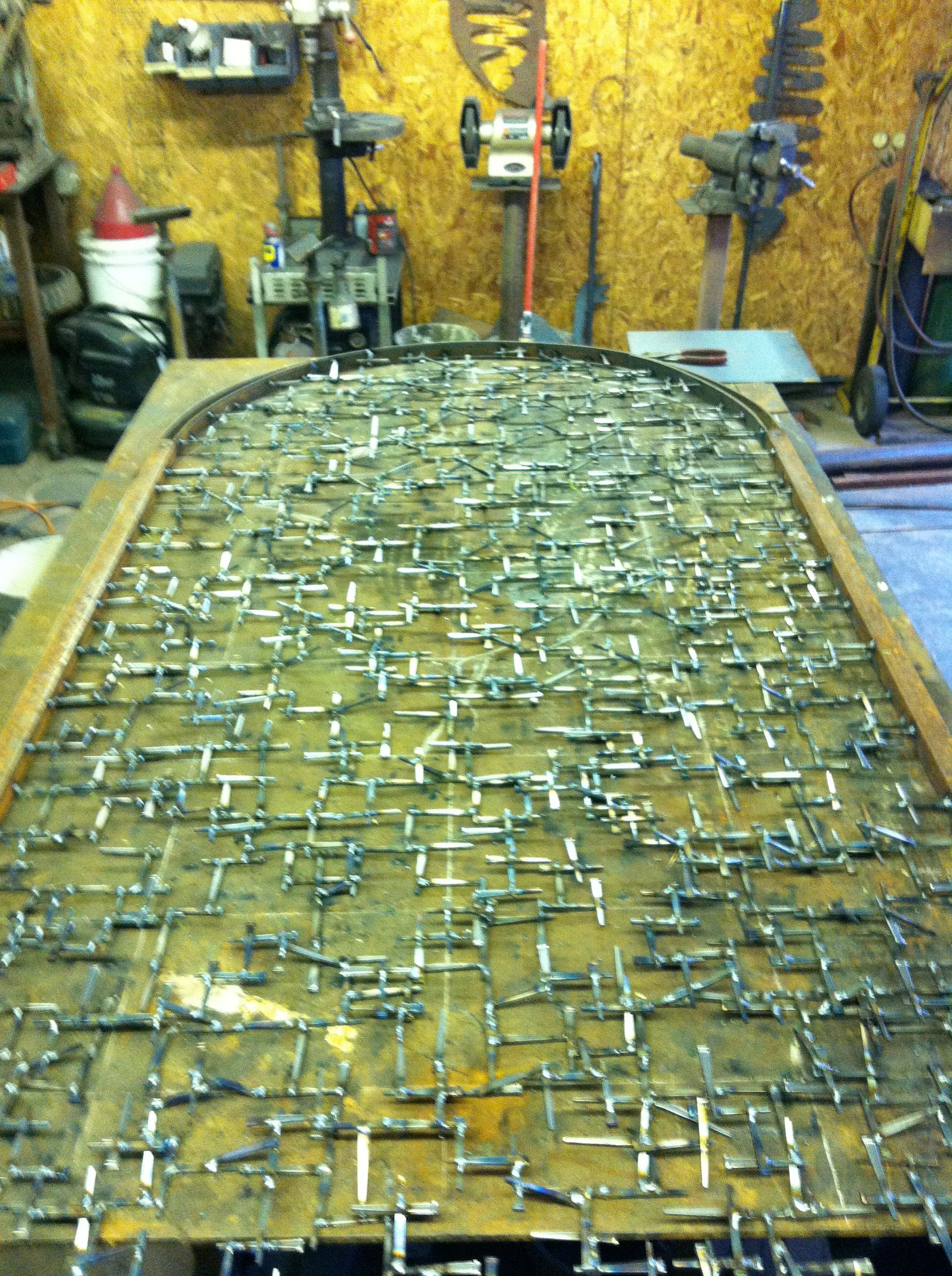 A long arched steel frame lying on a shop table, its inside filled with short nails welded crosswise, with tools and a workbench behind.