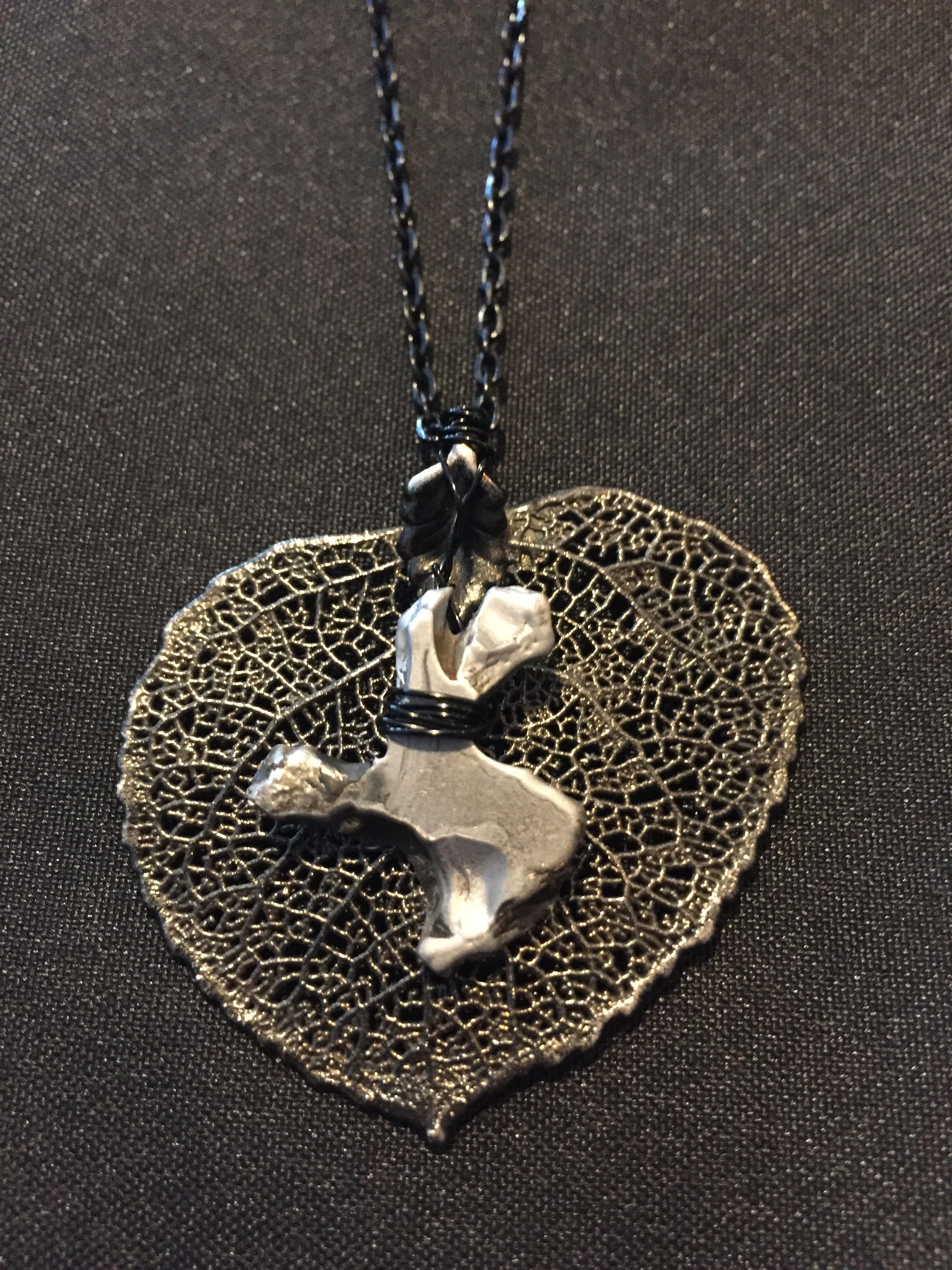 A bronze leaf-skeleton pendant with a silver cast piece laid across it, on a dark chain.