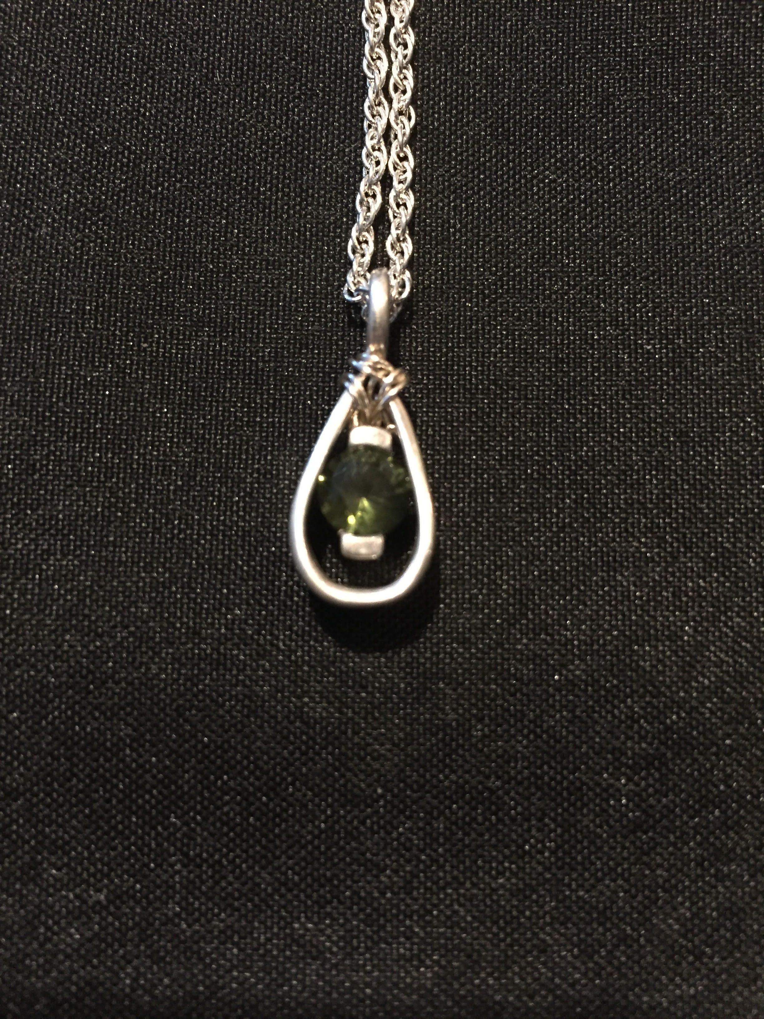 A small silver teardrop frame holding a green stone, on a silver chain against black cloth.