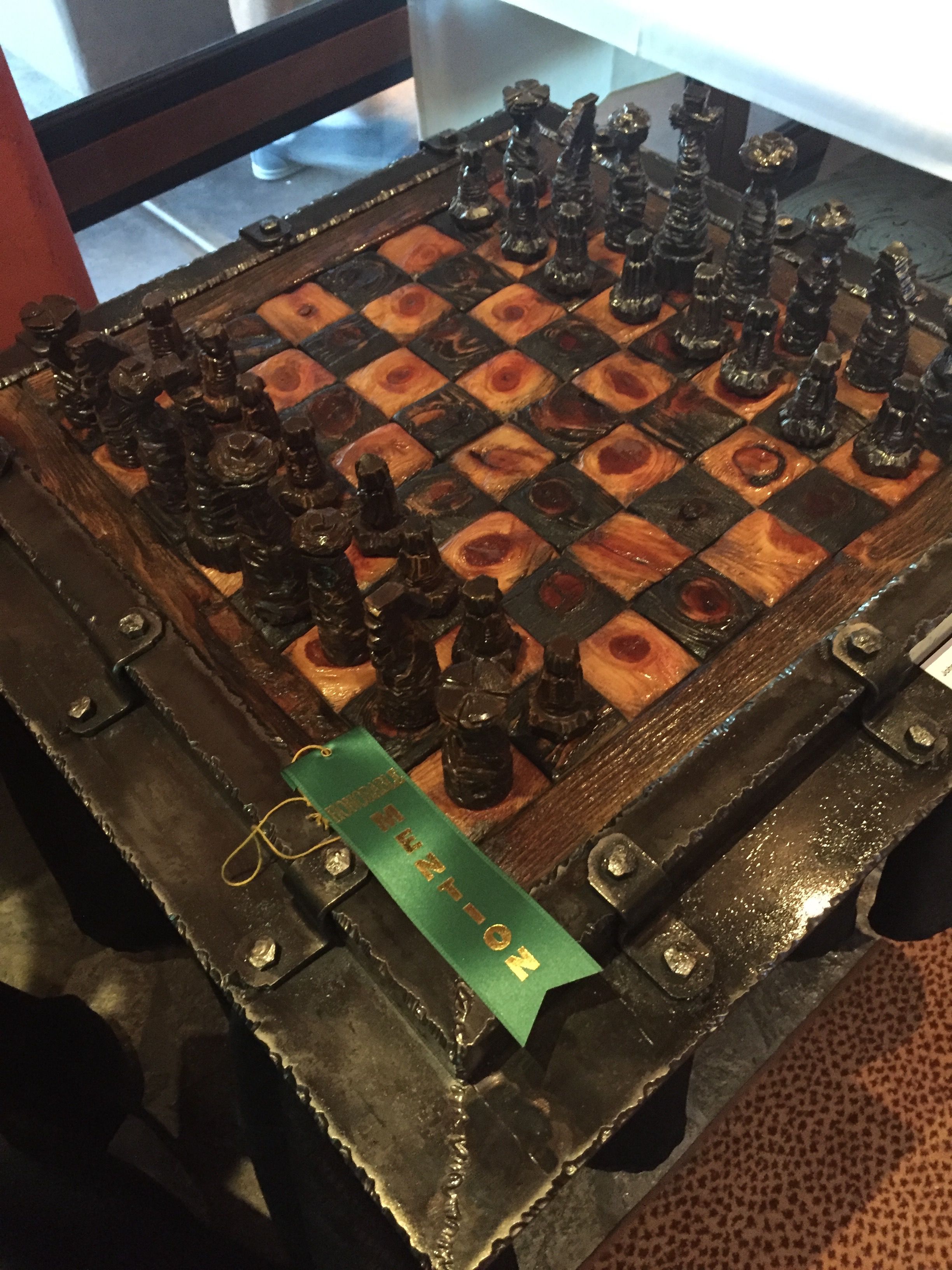 A chess set on a board of dark and light squares in a riveted steel frame, with a green ribbon lying across one corner.