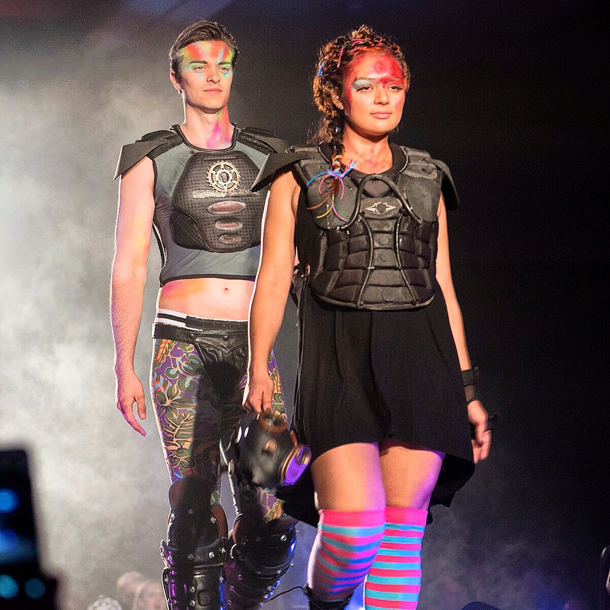 A man and a woman walk a runway in black armored vests; she wears striped knee socks and a short black skirt.