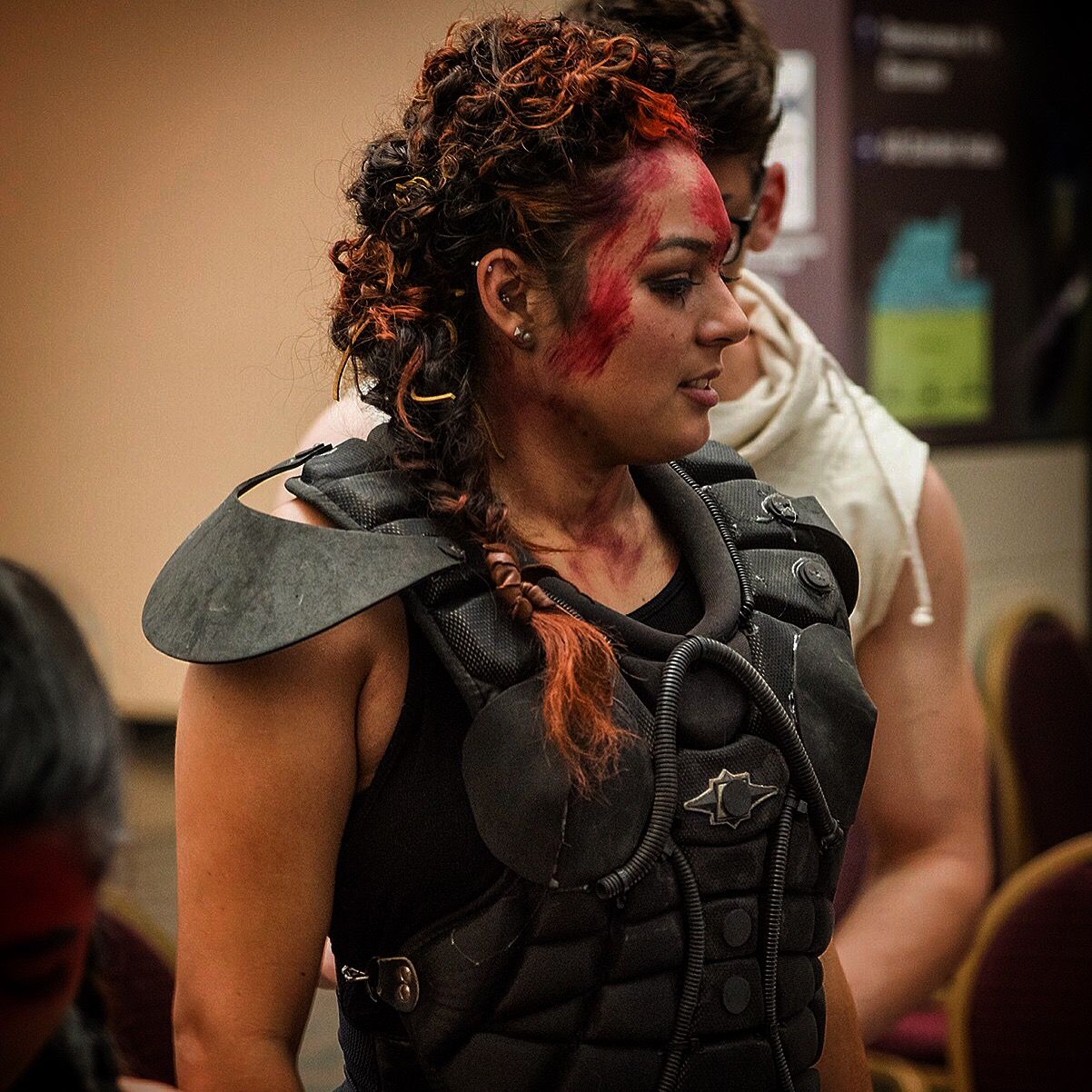 A woman with braided hair and red face paint wears a black shoulder guard over an armored vest.