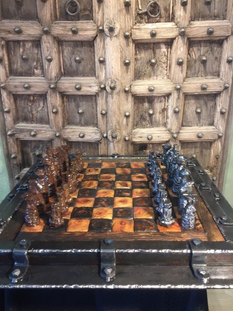 A chess board in a steel frame with bronze pieces on one side and dark blue-silver pieces on the other, in front of a studded wooden door.