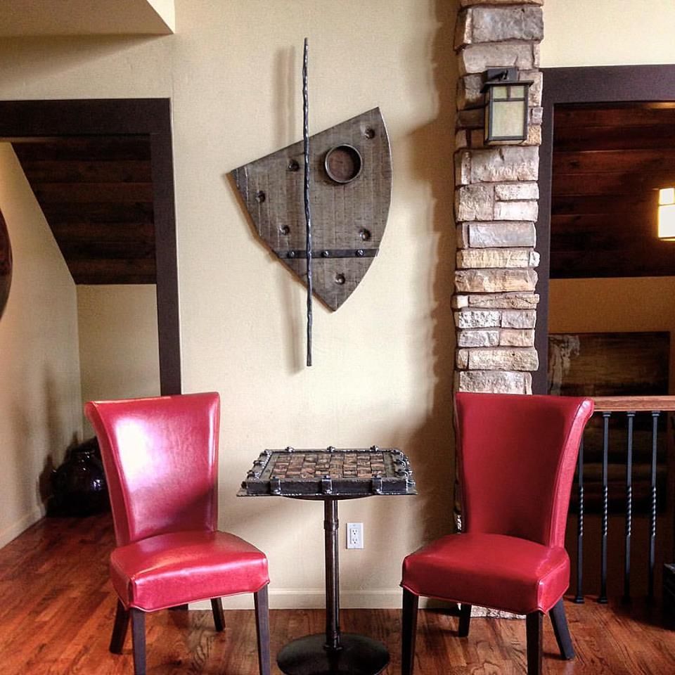 A small chess table between two red leather chairs, below a shield-shaped wall piece and a spear, beside a stone column.