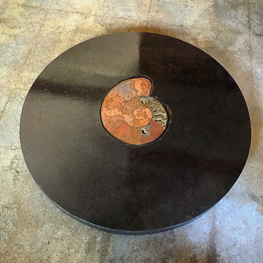 A round dark steel lazy susan with a polished ammonite fossil set near its center, on a stone floor.