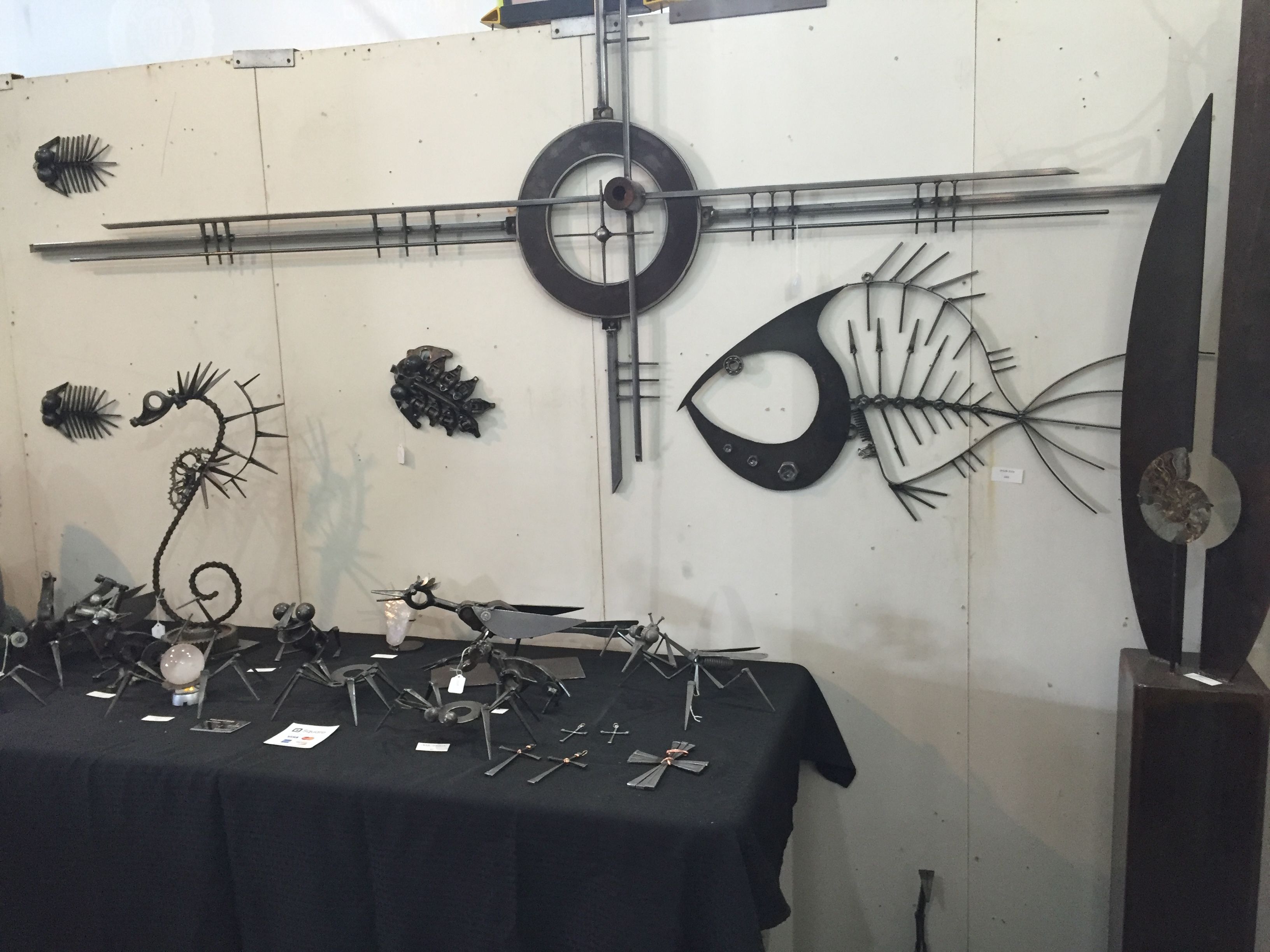 A black table covered with small steel creatures and a seahorse, below a long horizontal wall piece with a ring at its center and a fish skeleton.