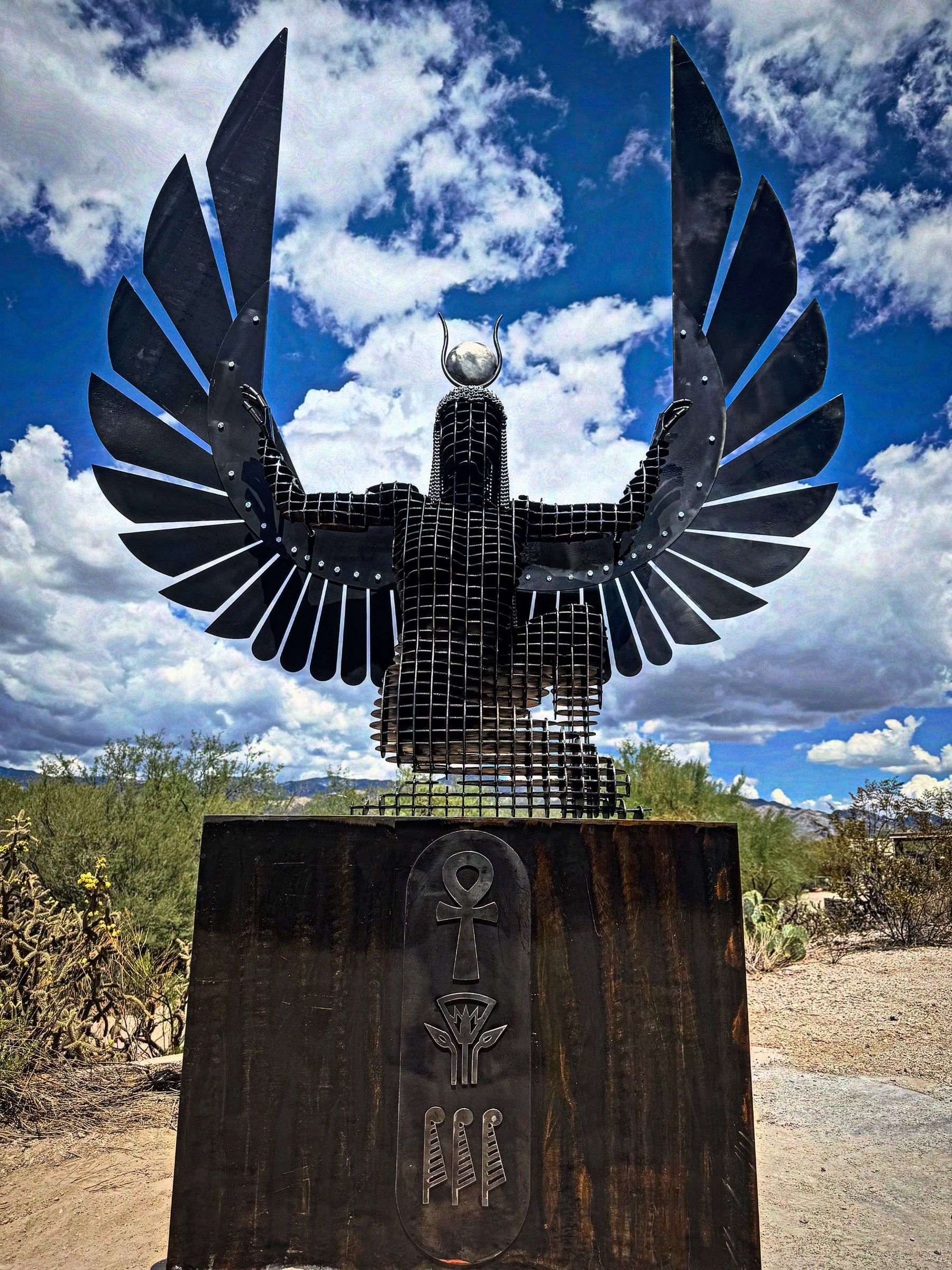 A winged figure of steel slices with both wings spread wide and arms raised, on a tall carved wooden base in the desert.