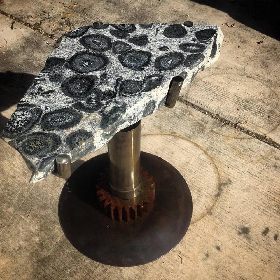 A slab of gray granite patterned with dark rings, on a single steel post with a gear at its foot over a round steel base.