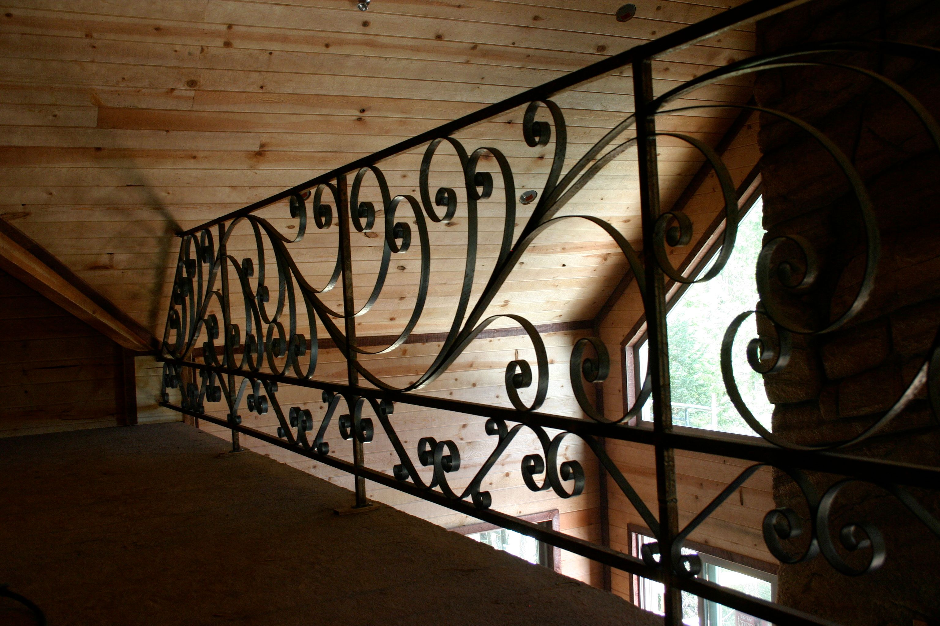 A black steel railing of scrolls and curls running along a loft, against wood paneling and a window.