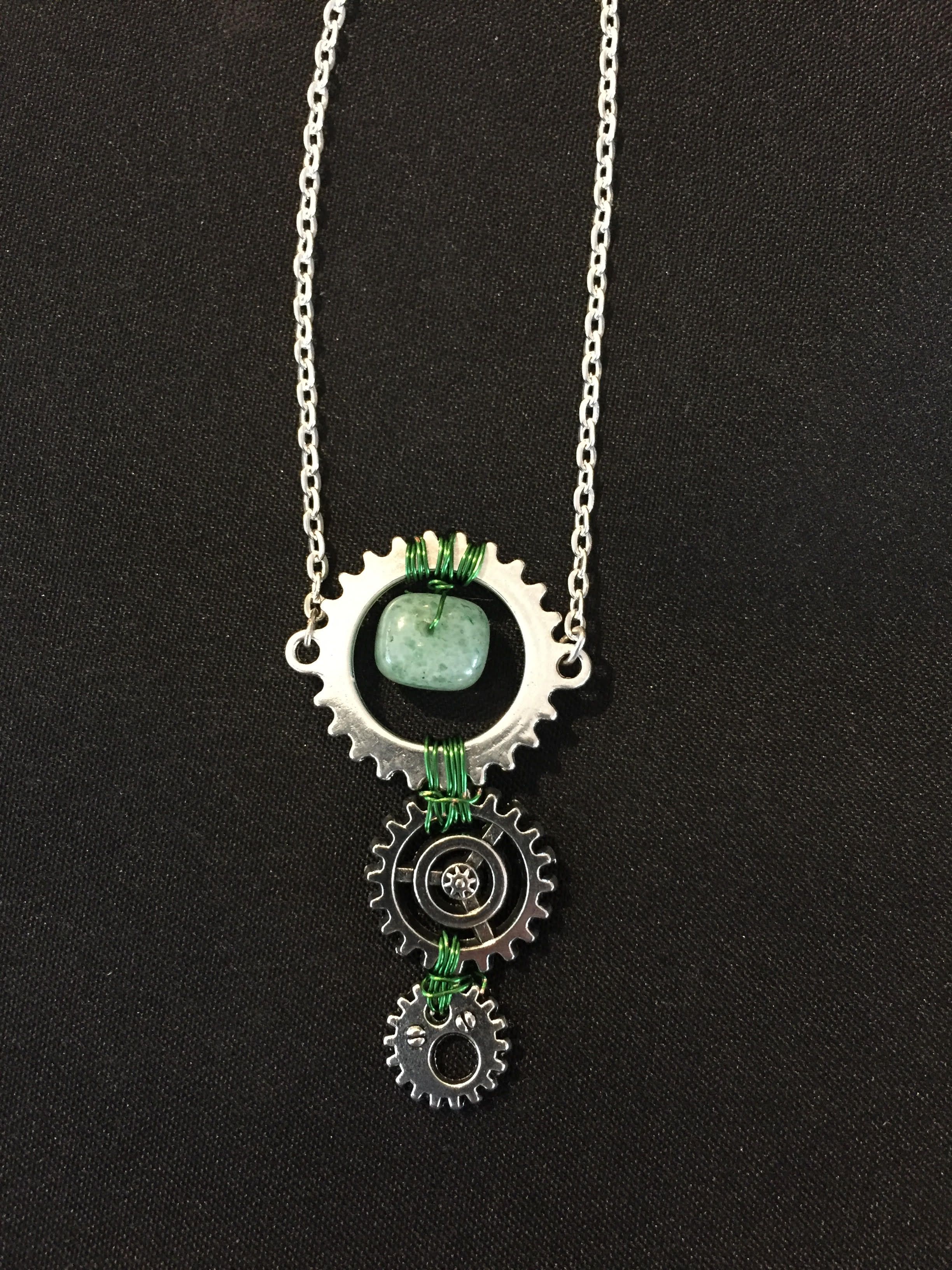 A silver chain holding a gear with a green stone at its center and two smaller dark gears hanging below.