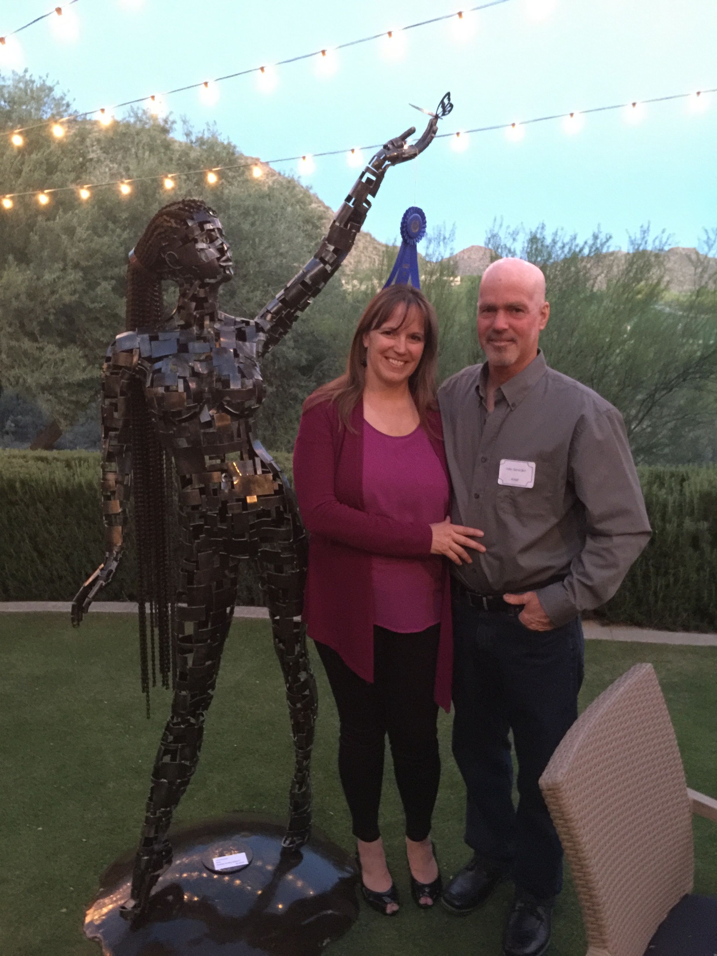 A man and a woman stand on a lawn beside the steel figure of Serenity, under string lights, with hills behind them.