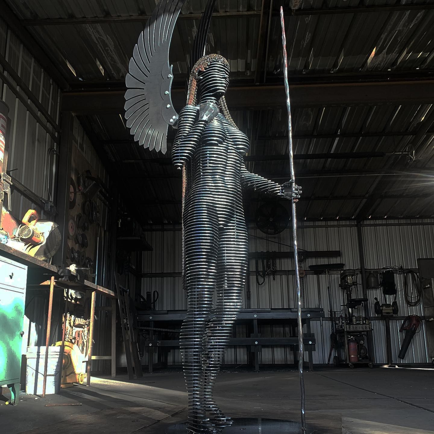 A life-size angel of stacked steel slices stands in a metal workshop, a polished wing curving above her shoulder and a tall staff in one hand.