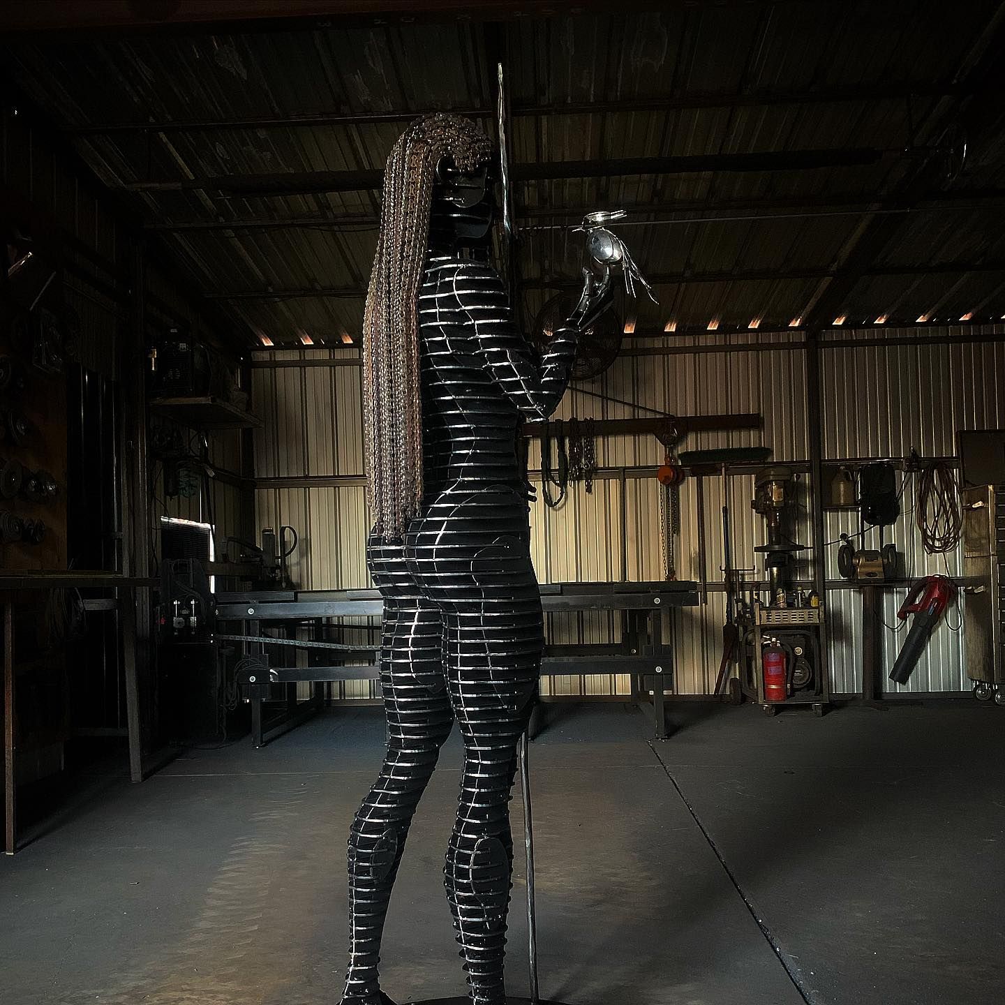 A standing figure of stacked steel slices with long braided hair, a bird perched on her raised hand and a staff beside her, in a workshop.