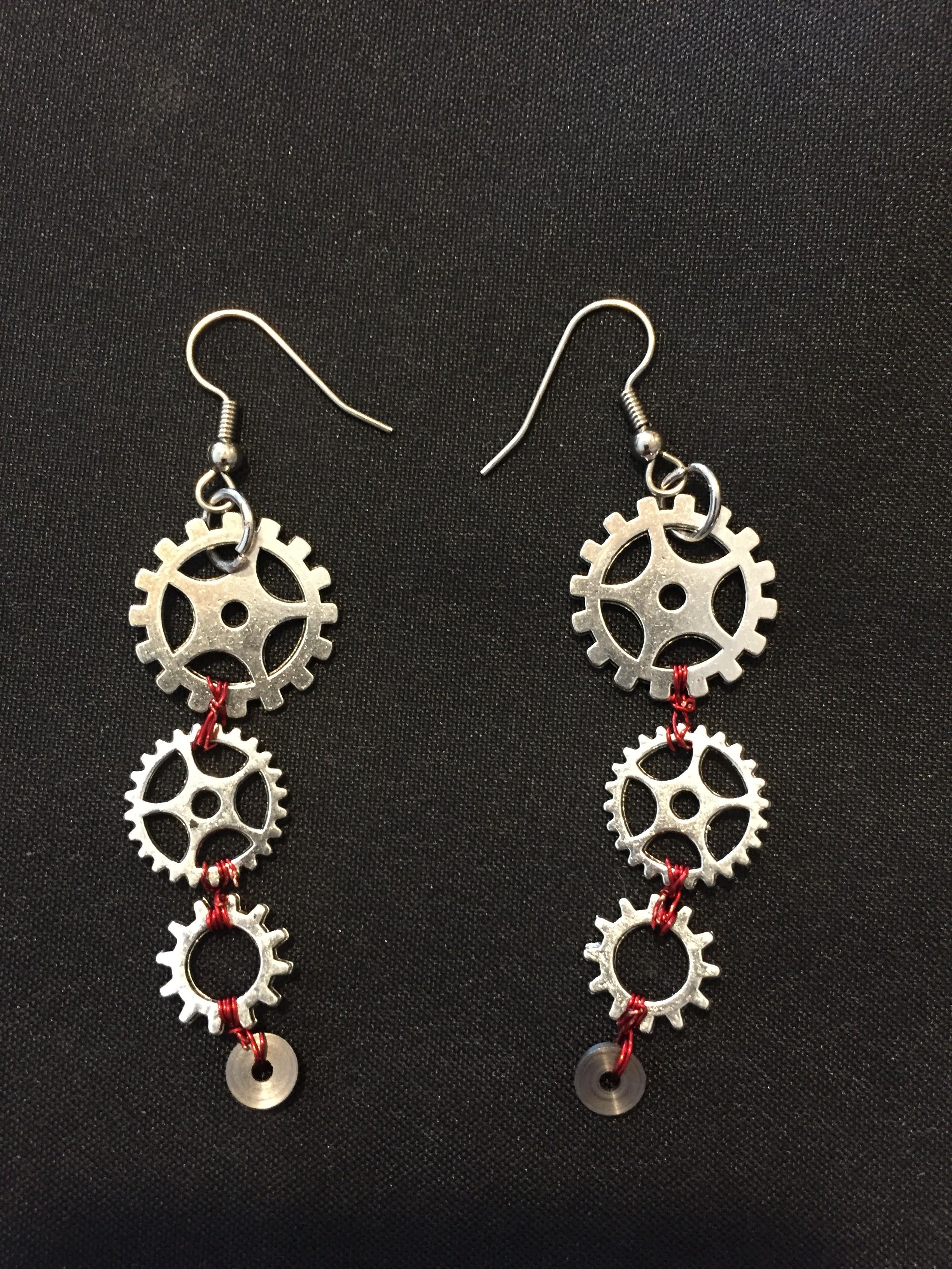A pair of earrings, each a column of three silver gears joined by small red beads, on black cloth.