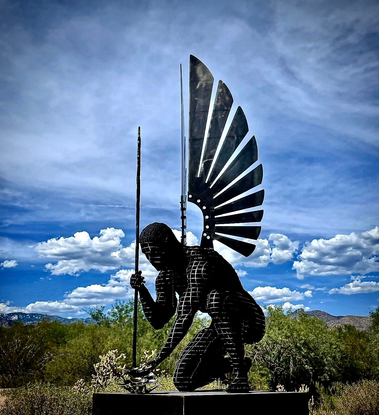 A dark angel of steel slices kneels with one hand reaching down, holding a tall spear, a fanned wing rising behind, under a blue sky with clouds.