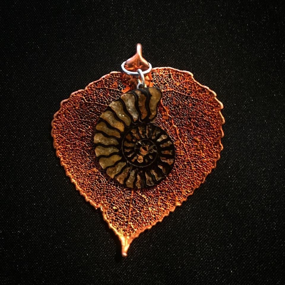 A copper leaf-skeleton pendant with a small ammonite fossil set on it, against black.