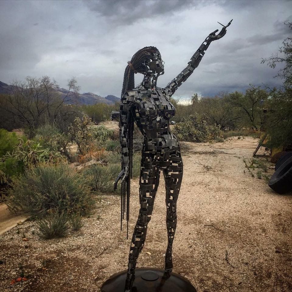 The steel figure of Serenity standing in the desert with one arm raised, long chain-like strands falling from her head, under a cloudy sky.