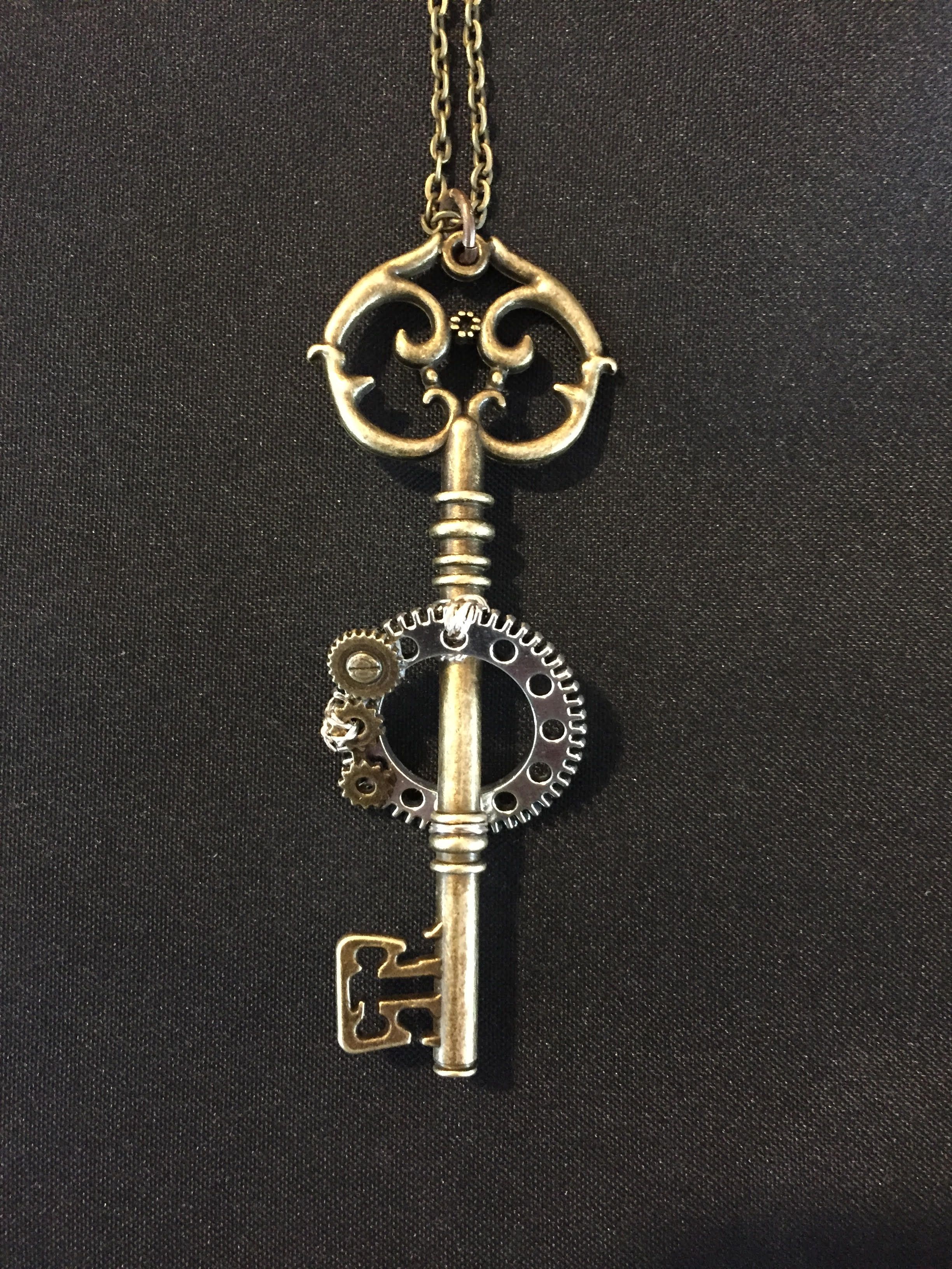 An ornate brass key pendant with a small gear set at its center, on a gold chain against black cloth.