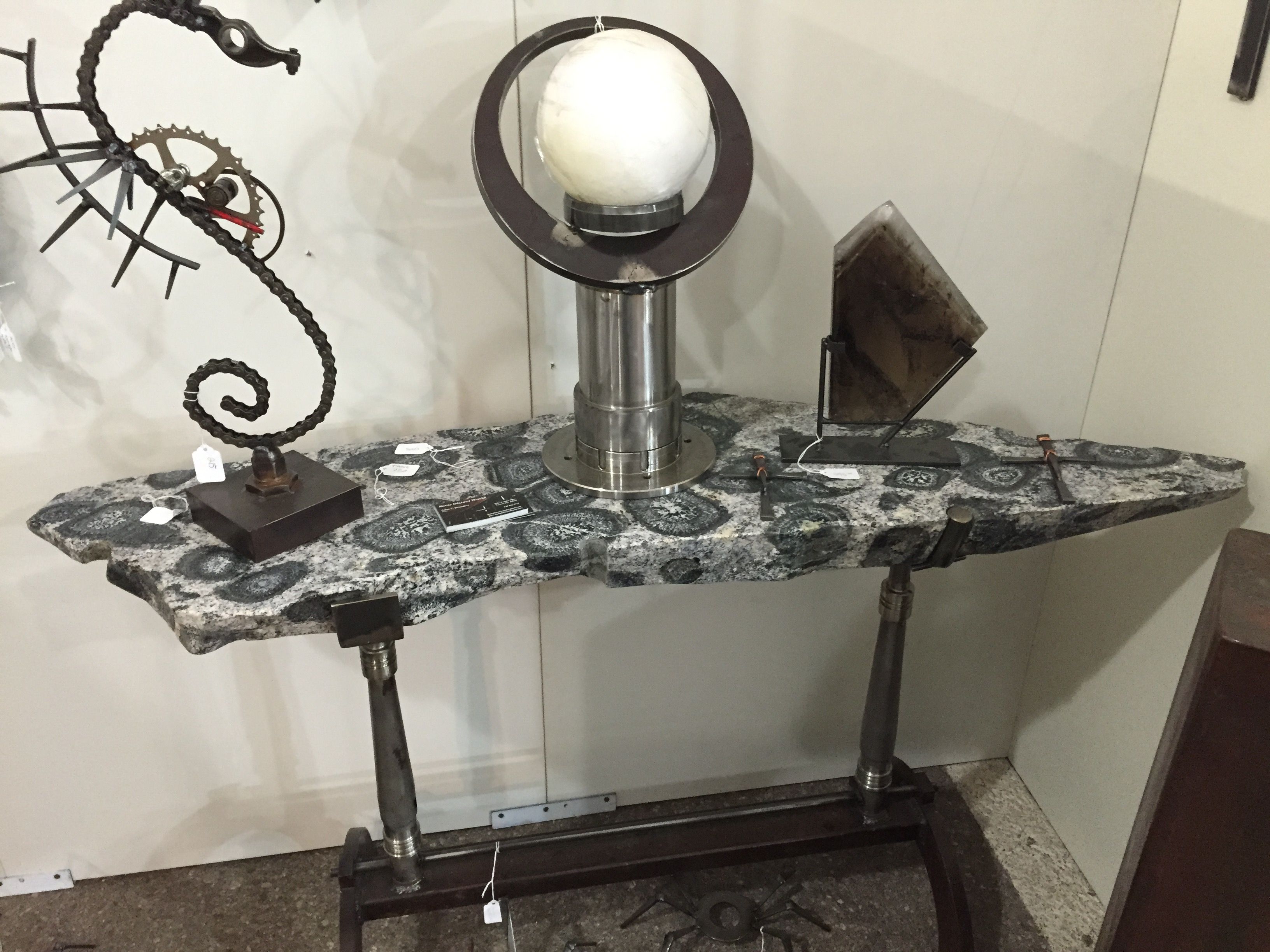 A console table with a gray ringed-stone top on steel legs, holding a lamp with a white globe in a steel ring, a steel seahorse at the left and a dark wall piece at the right.