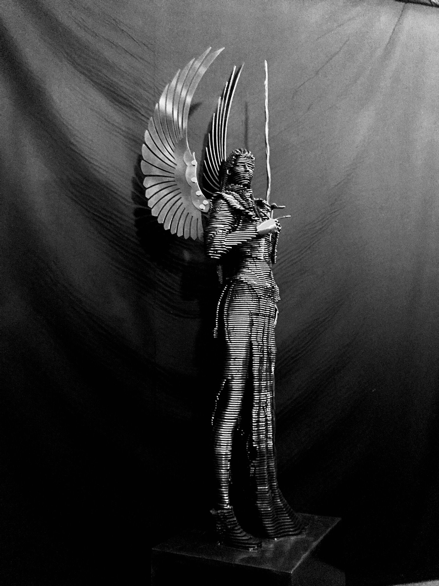 A standing steel angel built of thin horizontal slices, polished wings raised behind her and a staff in hand, on a low base against a dark backdrop.