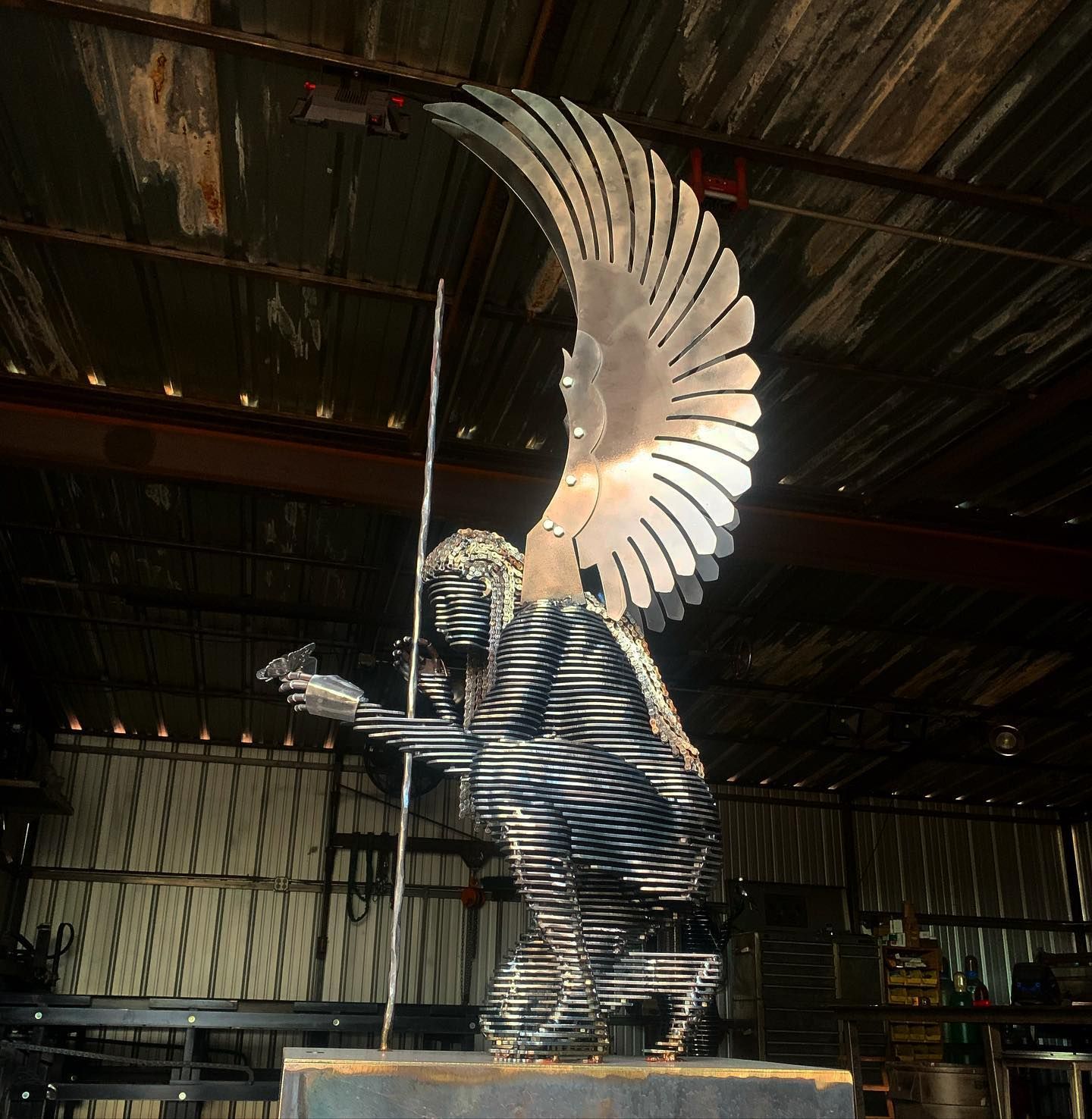 A kneeling angel of steel slices with a large polished wing and a spear, in a workshop.