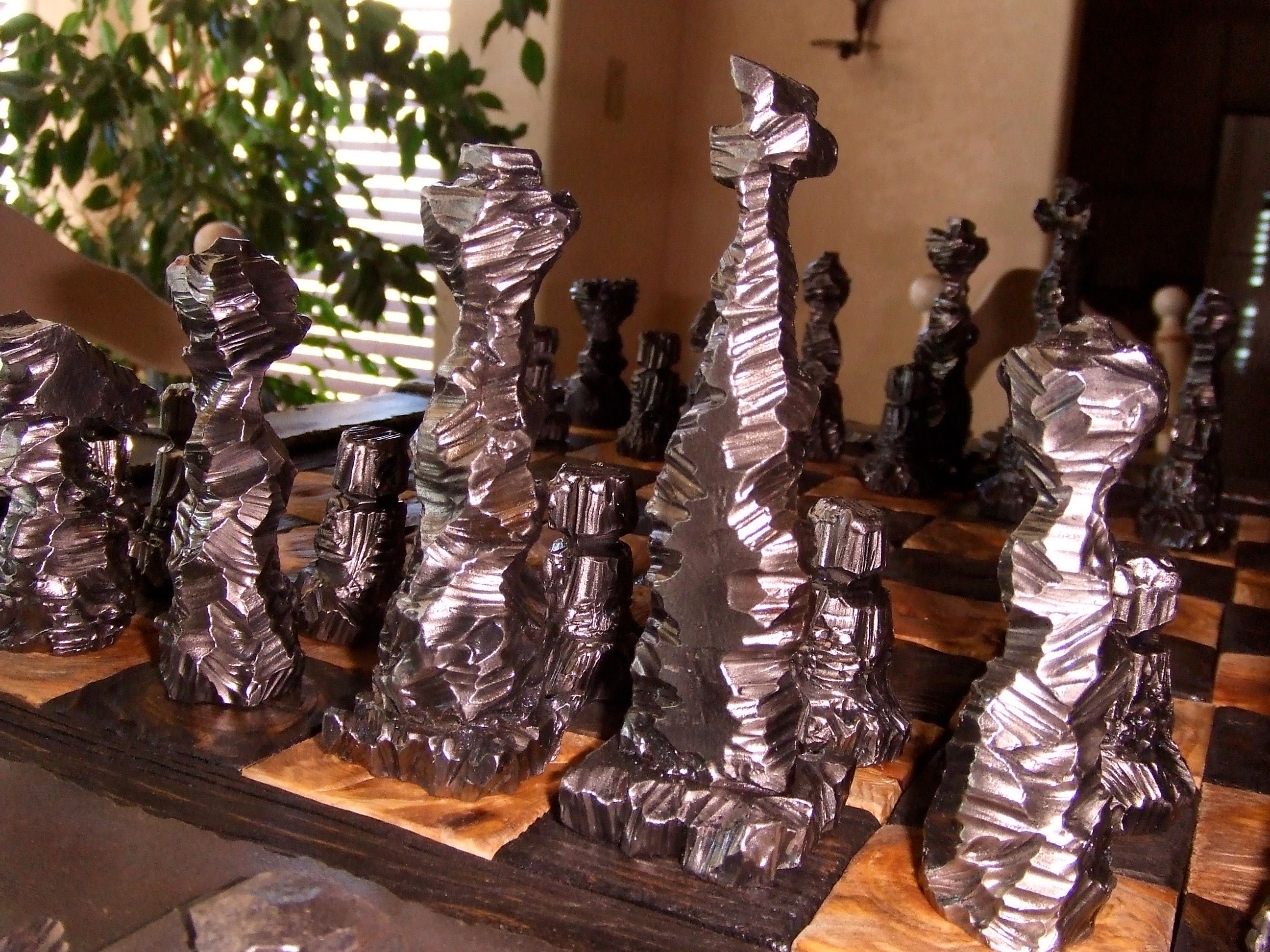 Tall bronze and silver chess pieces with twisted, folded surfaces stand on the board in front of a window and plants.