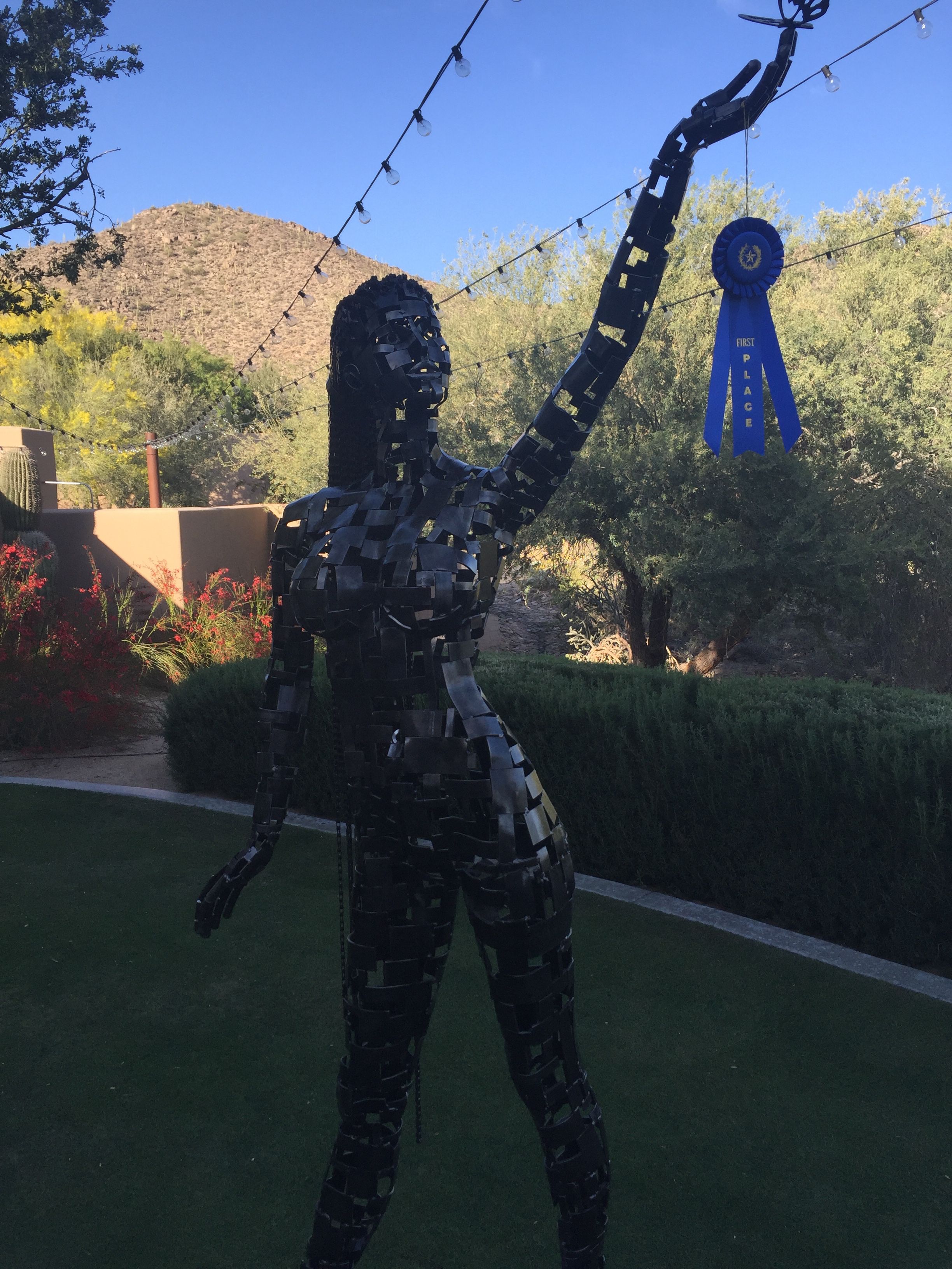 The steel figure of Serenity in daylight on a lawn, a blue ribbon hanging from her raised hand, a mountain and trees behind her.