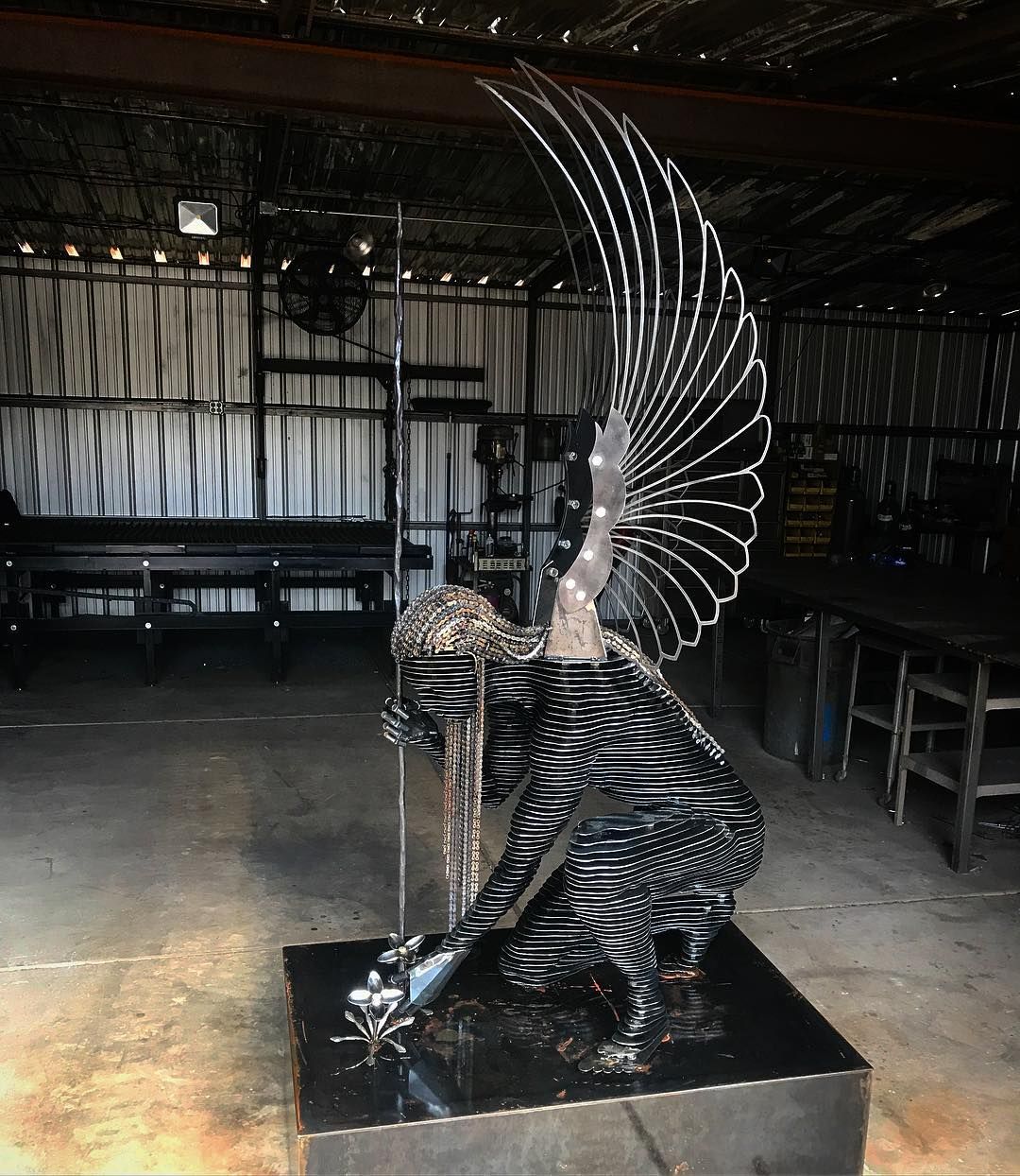 A kneeling angel with a tall fanned wing and a spear, reaching down toward the top of a square steel base, on a workshop floor.