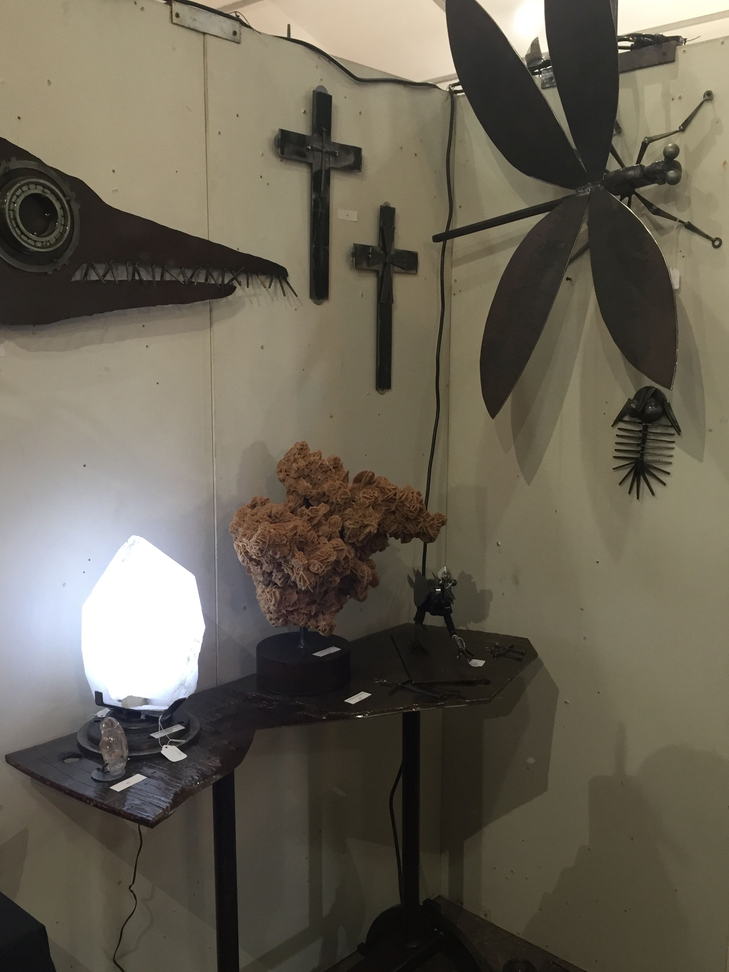 A white booth corner with a large steel dragonfly, two crosses and a fish-head wall piece, and a glowing lamp on a small table.