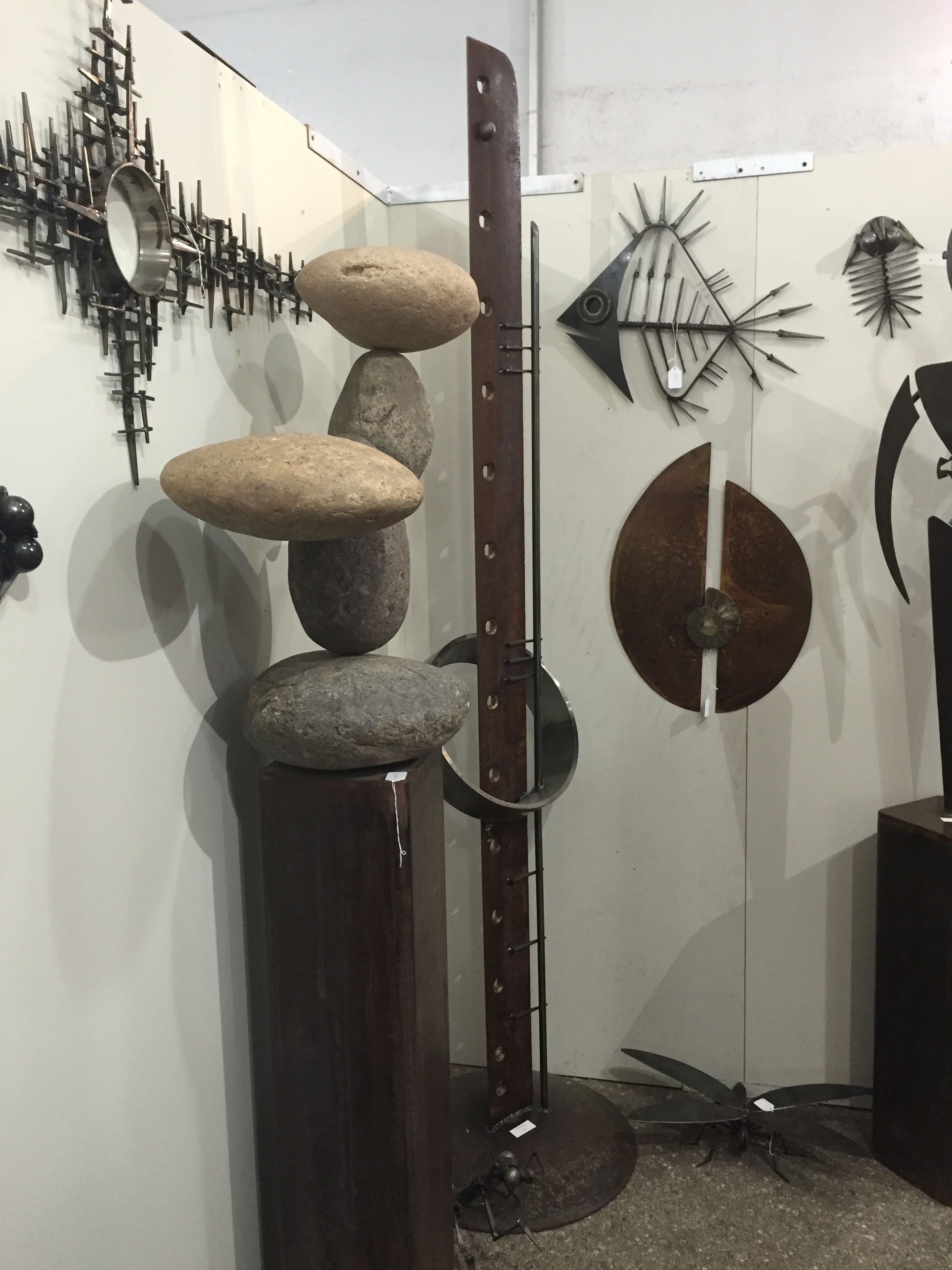 Smooth stones balanced on top of a tall rusted post, beside a round wall disc and a fish skeleton on a white wall.