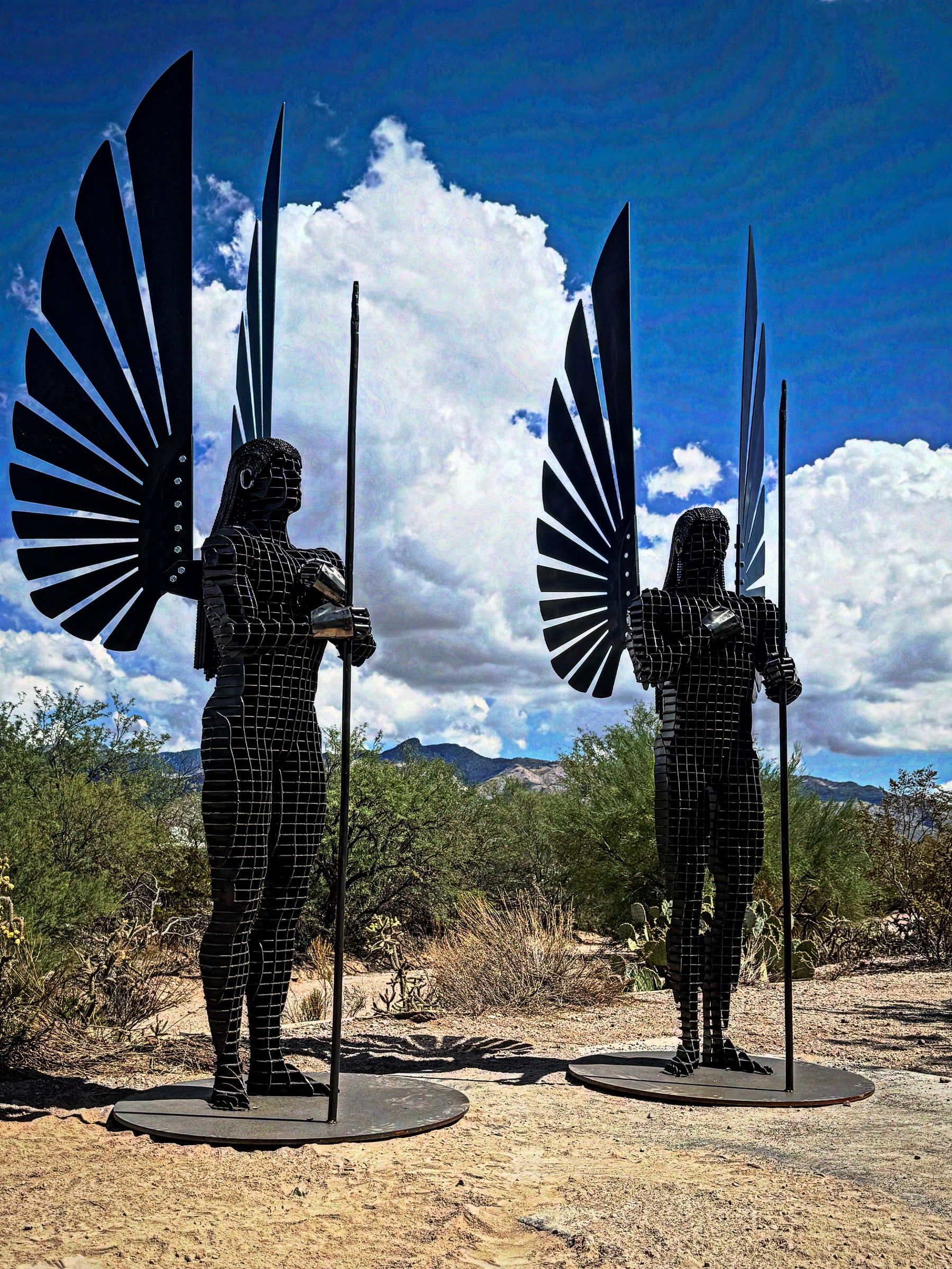 Two angels of steel slices stand on round bases in the desert, each with a fanned wing and a tall spear.
