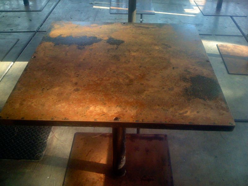 A square table with a mottled copper-colored top on a dark steel pedestal.
